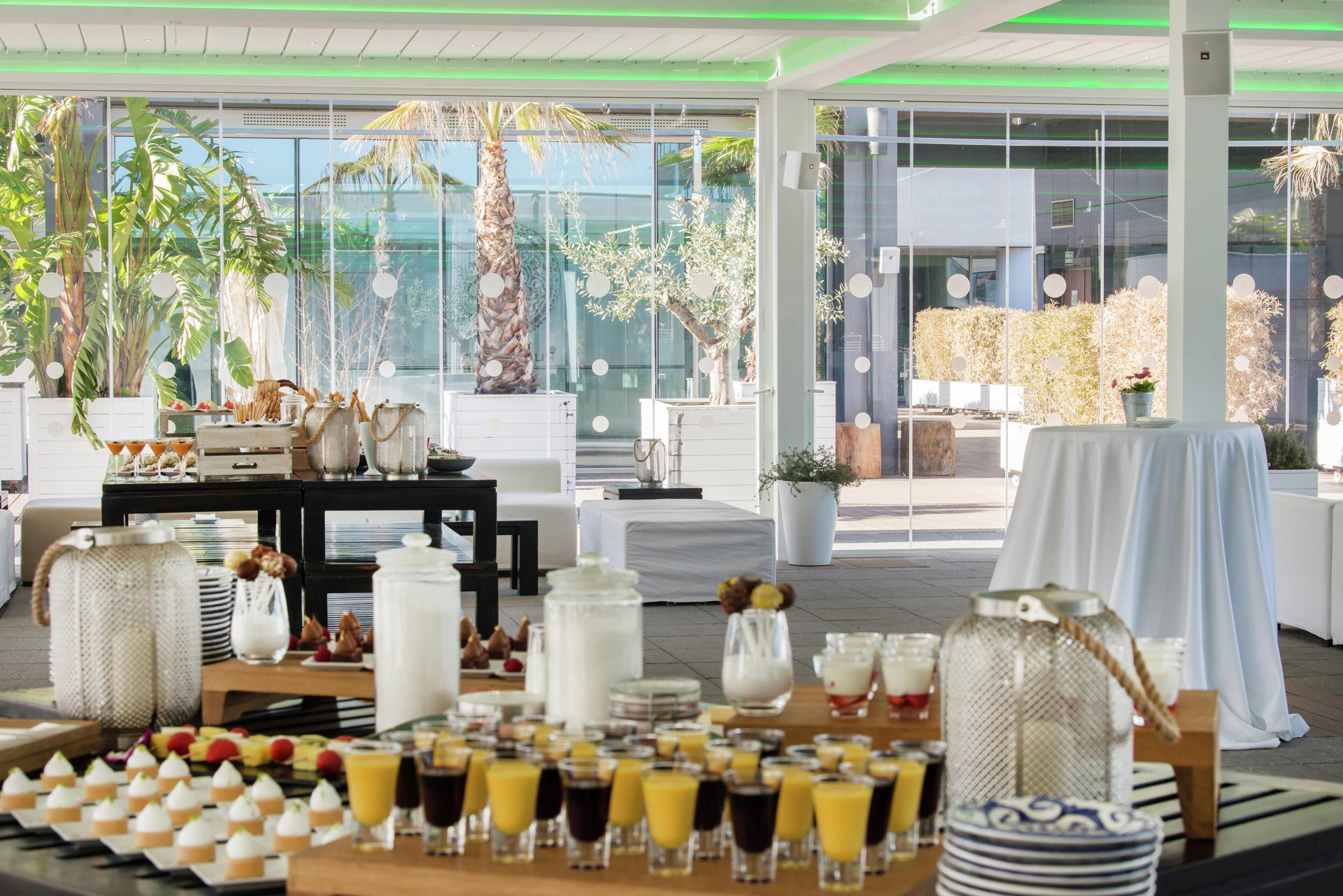 A breakfast buffet setup with various food items and drinks, surrounded by indoor plants and modern furniture.