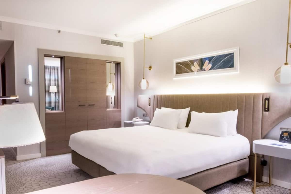 A modern hotel room with a large bed, a bedside table, a desk, and a round table.