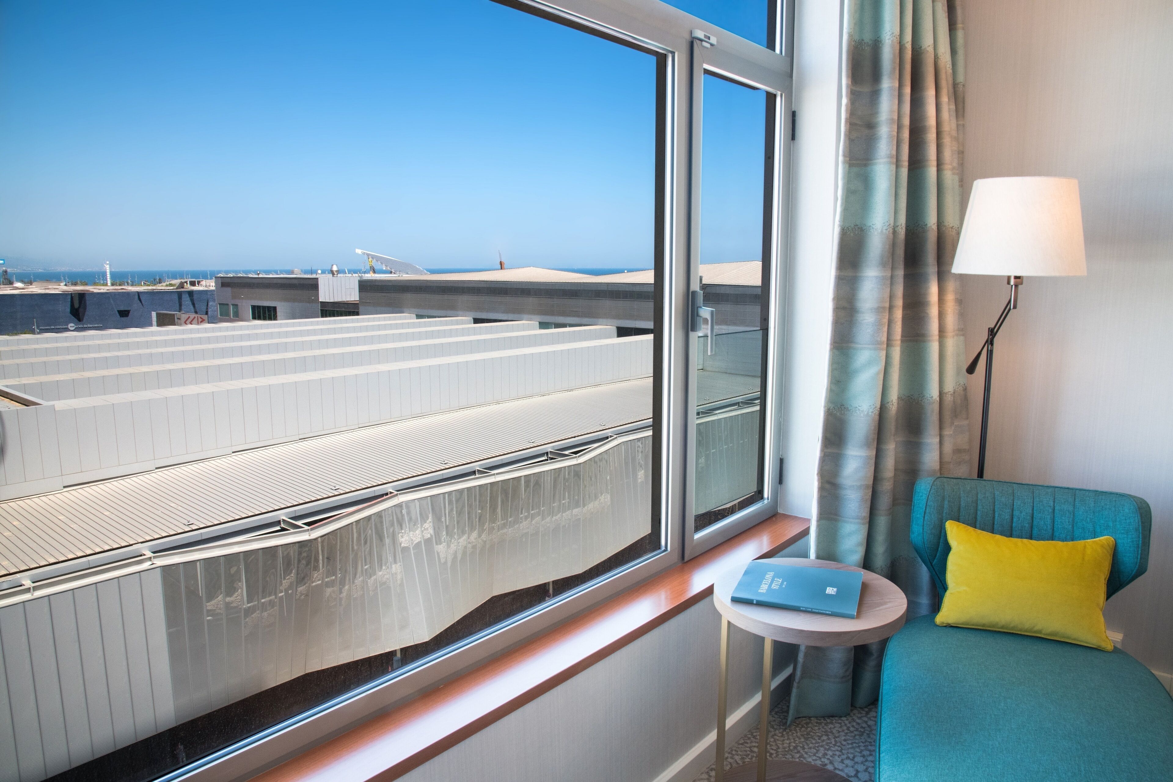 A hotel room with a large window offering a view of a coastal area, a small round table with a laptop, a teal armchair with a yellow pillow, and a floor lamp.