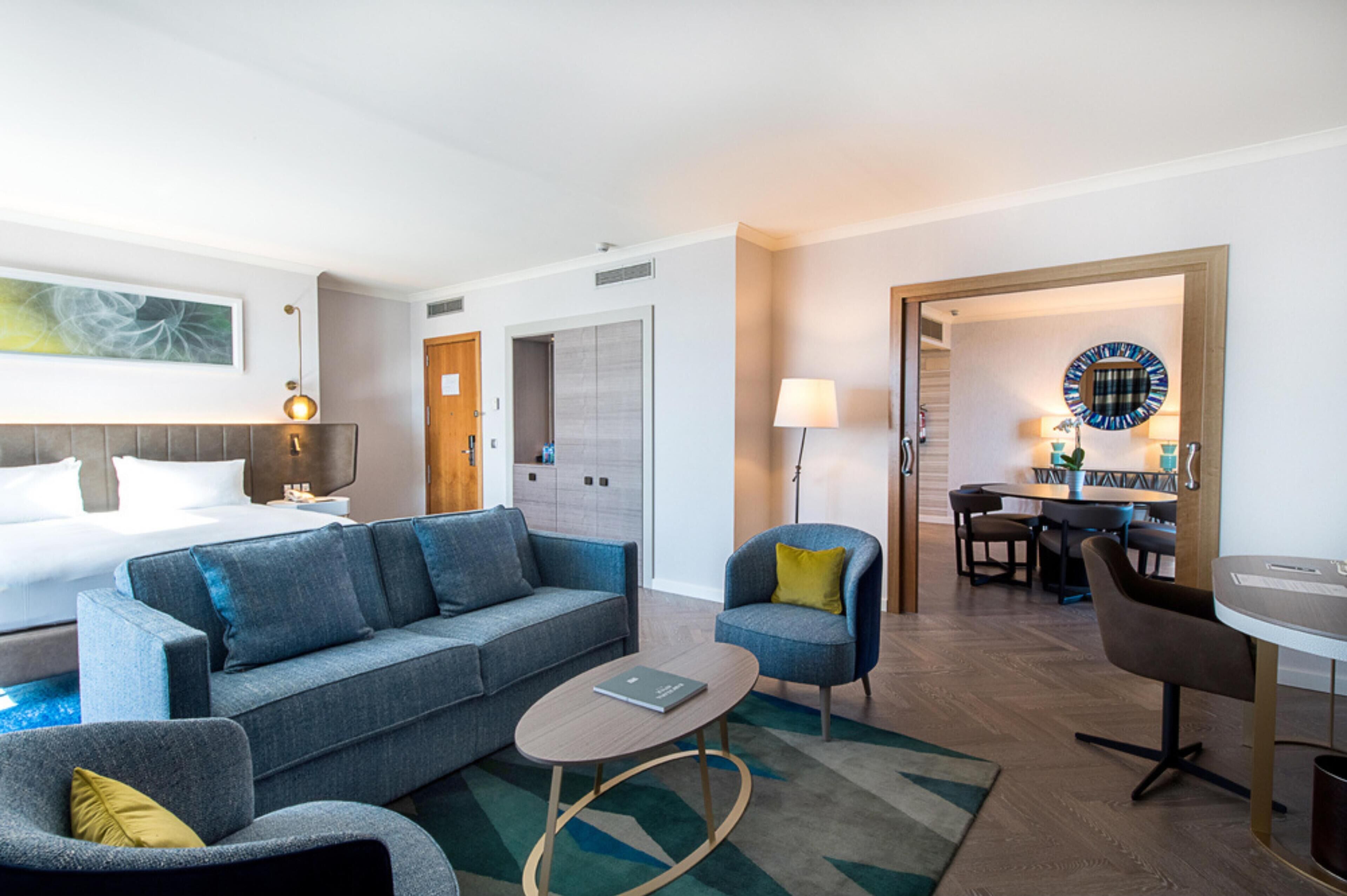 A hotel room with a bed, a grey sofa, a small armchair, a coffee table, a desk, and a dining area with a table and chairs.