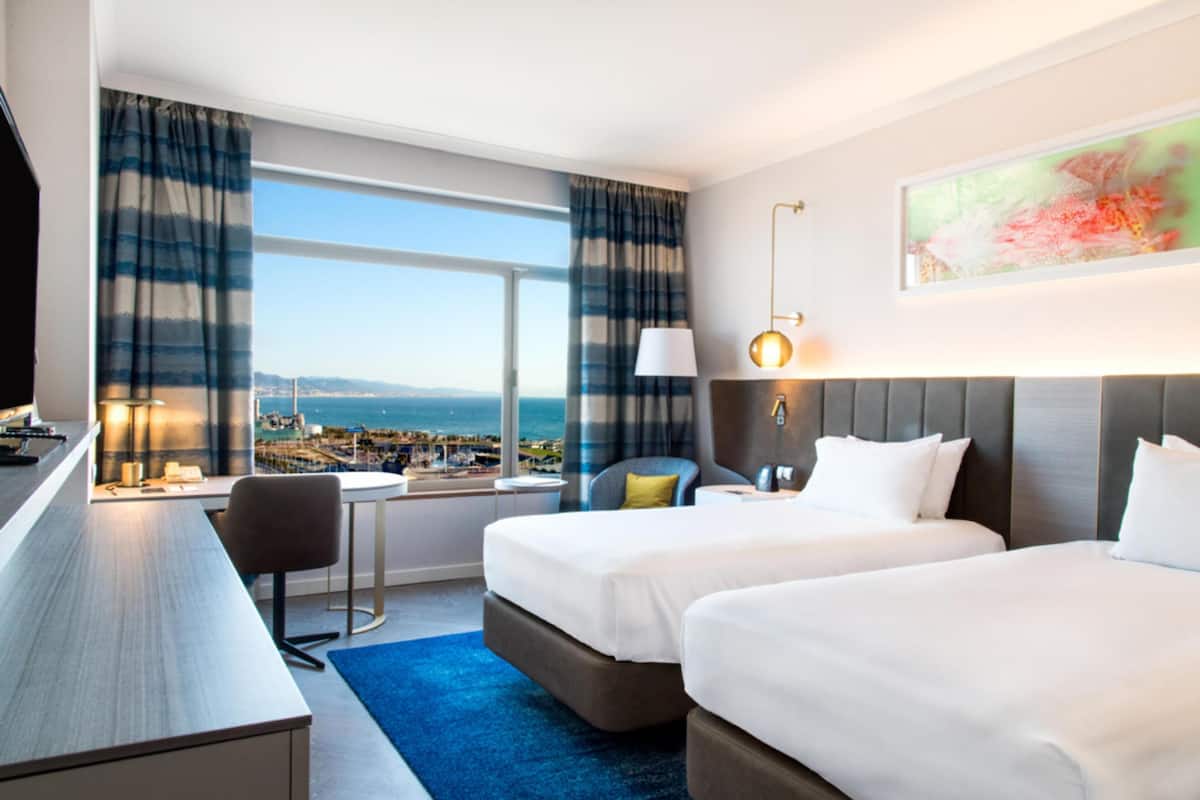 A hotel room with two beds, a desk, a chair, a lamp, and a view of the city and ocean.