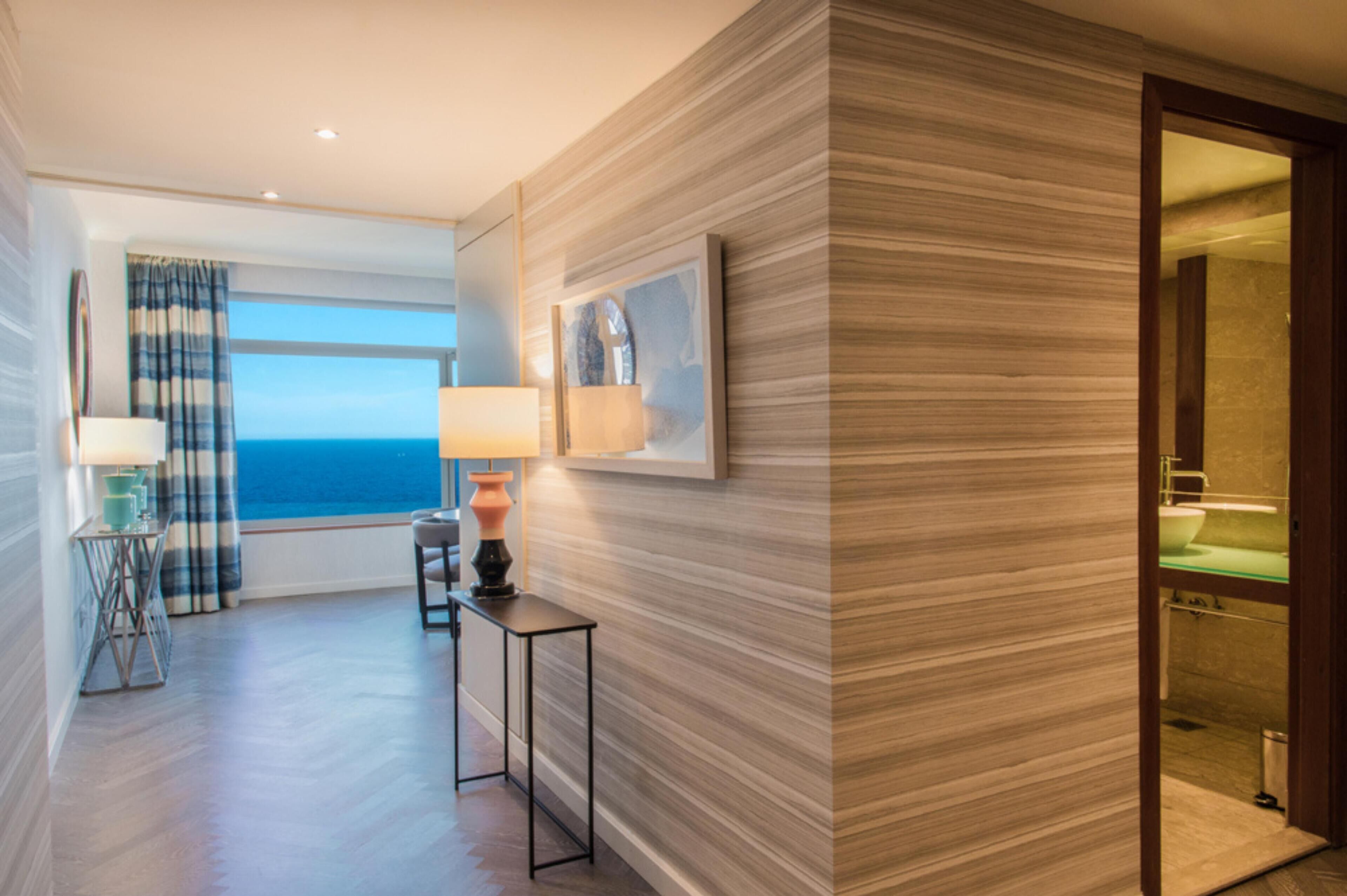 A hotel room with a view of the sea, a small table with a lamp, and a modern bathroom with a glass shower.