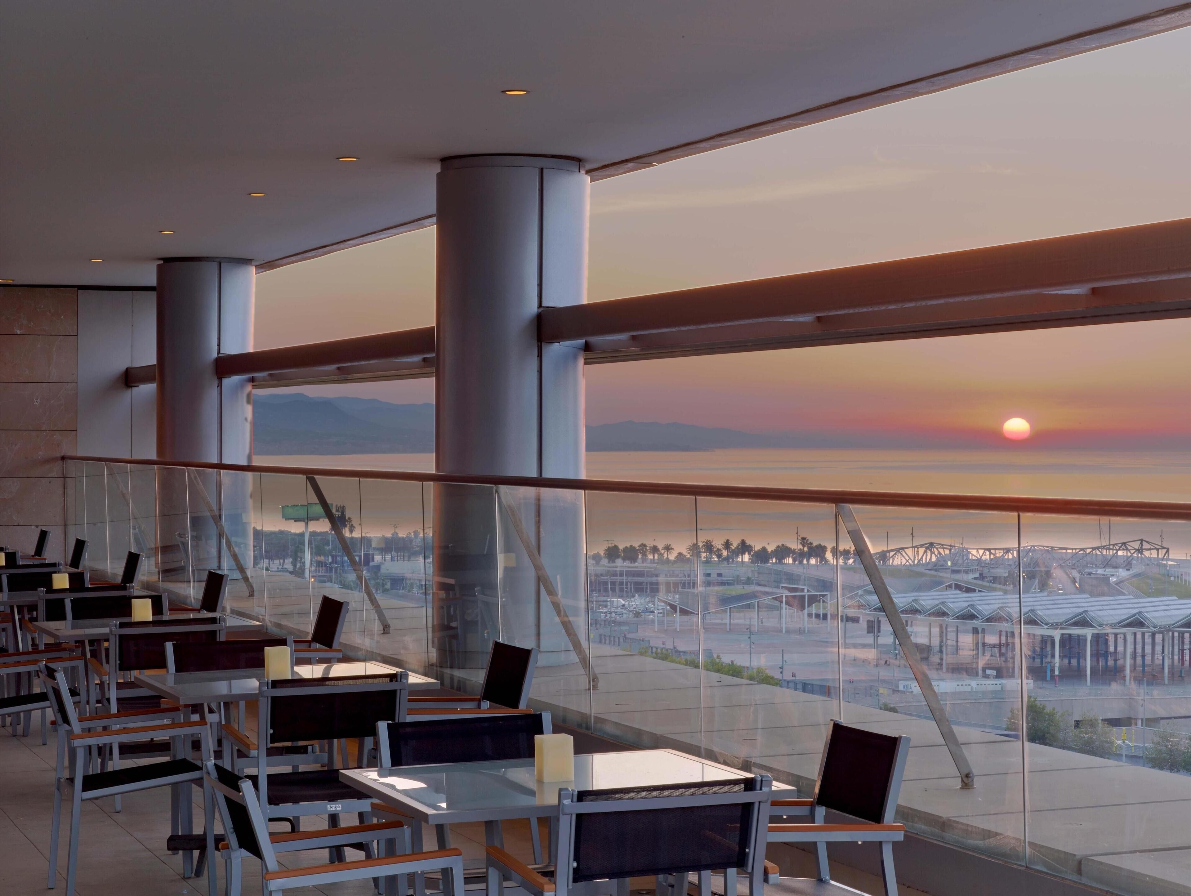 A modern restaurant with glass walls and a view of a sunset over a cityscape.