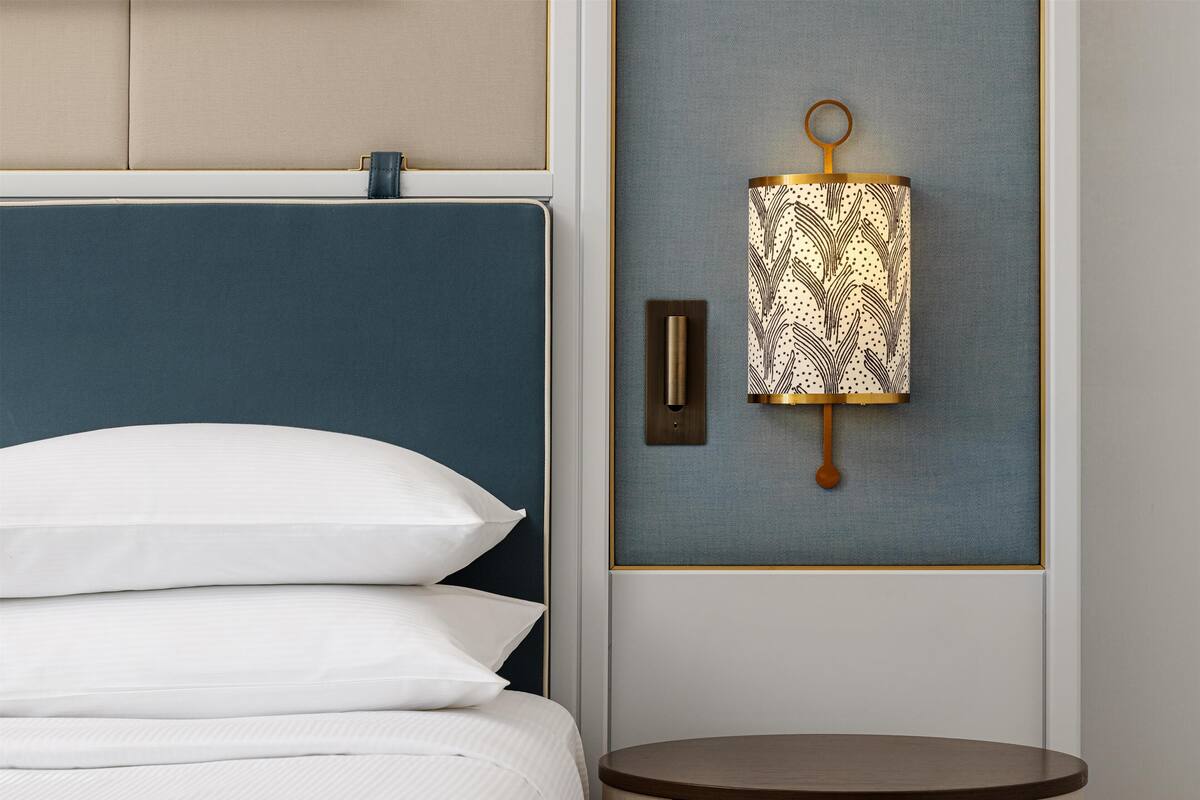 Een bed met witte lakens, een blauw hoofdbord en een rond nachtkastje van hout.