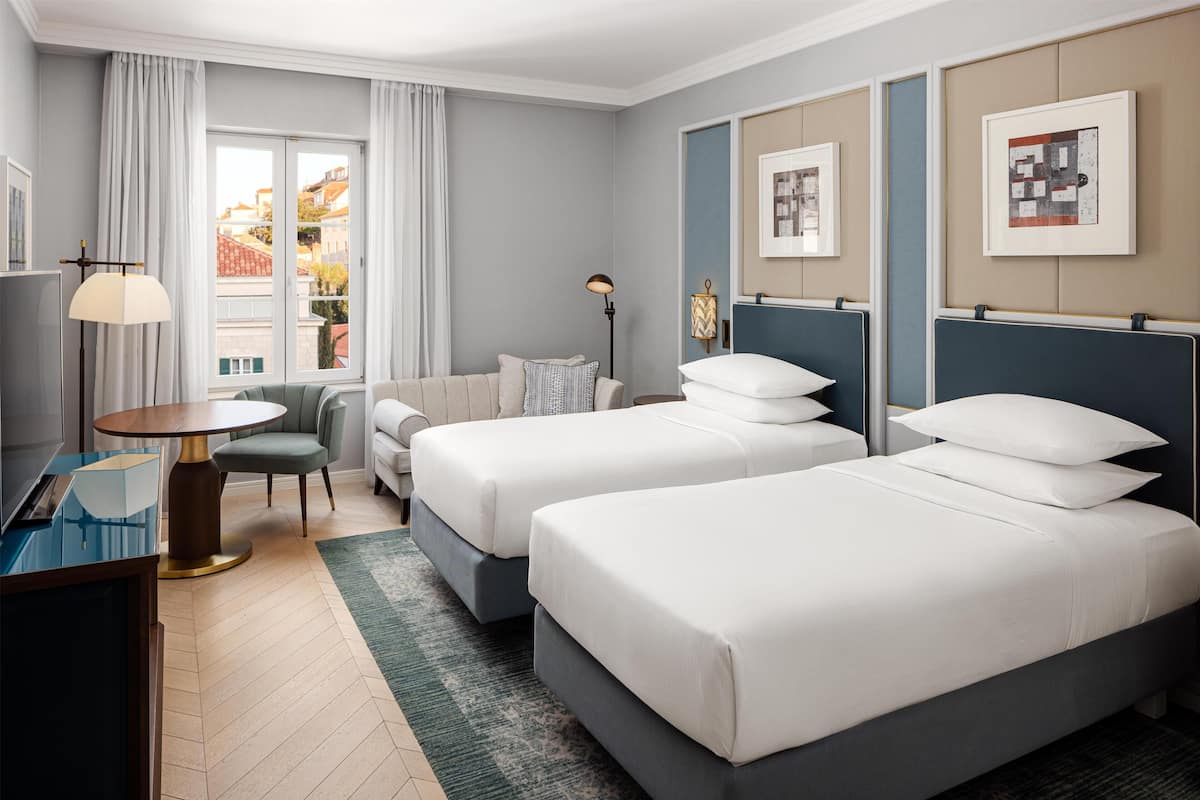 A hotel room with two beds, a desk, a chair, a lamp, and a view of buildings outside.