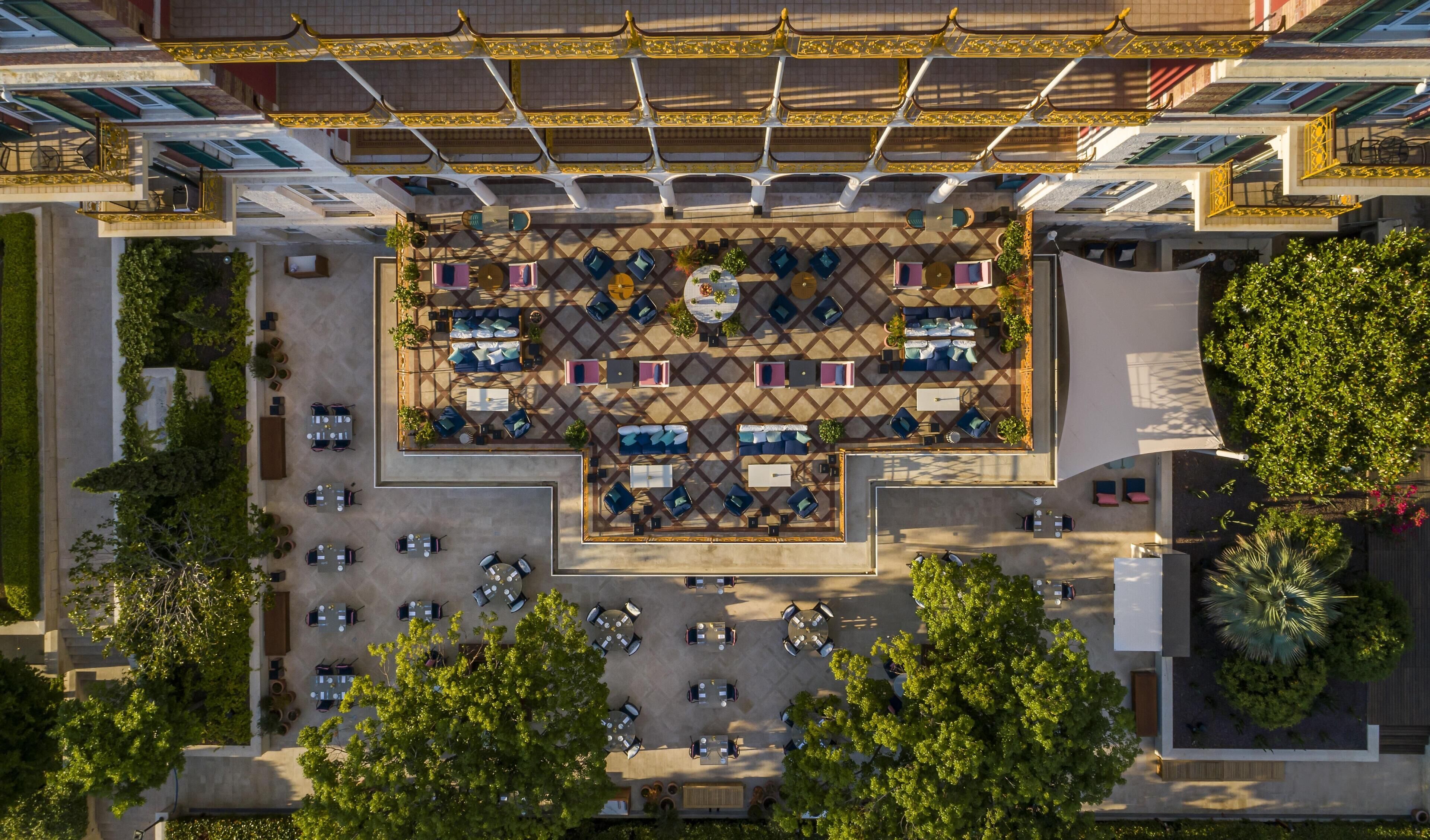 Vista aérea de un patio de hotel con mobiliario exterior, una fuente central y abundante vegetación.