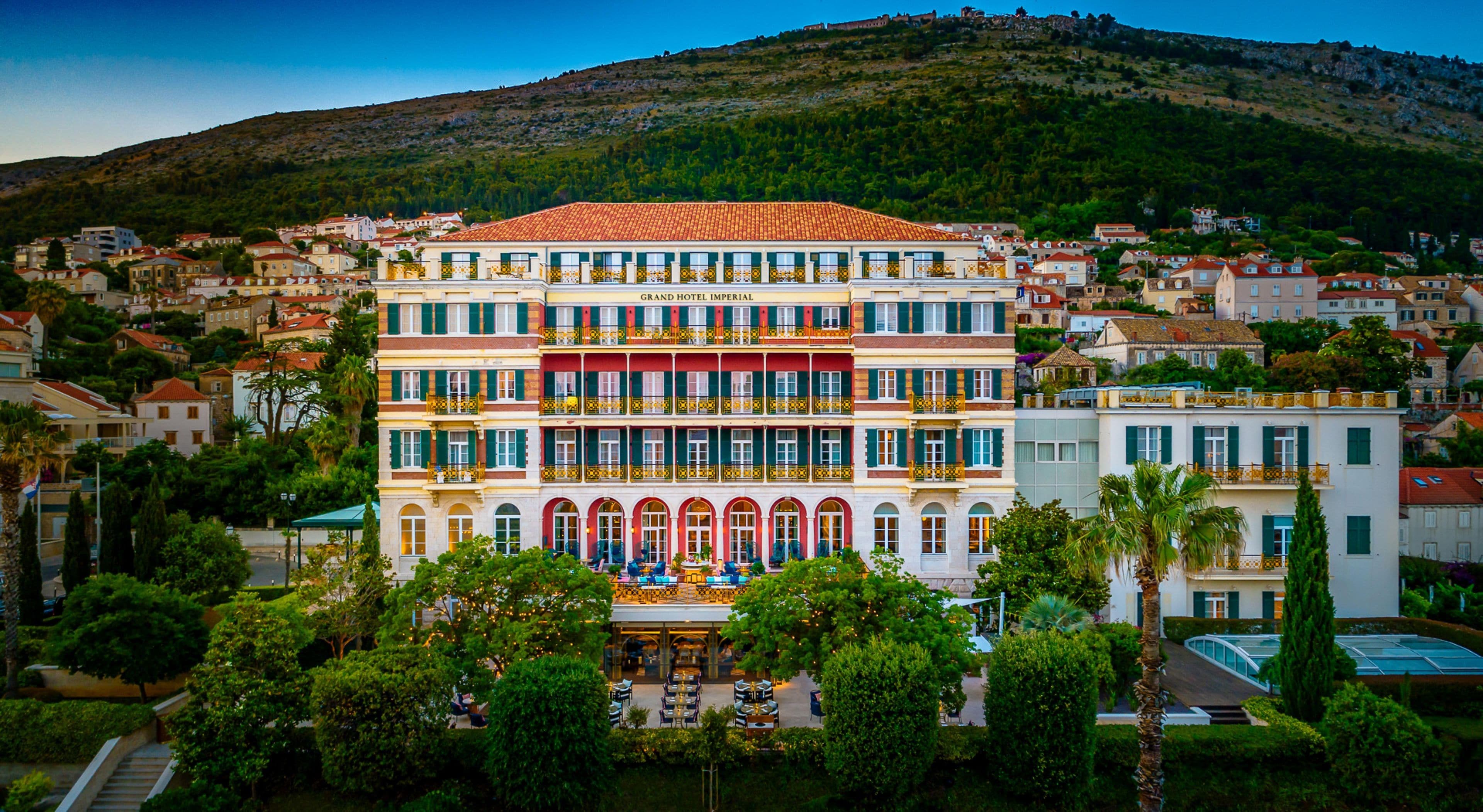 A multi-story hotel with a red-tiled roof, multiple balconies, and a garden area with outdoor seating.