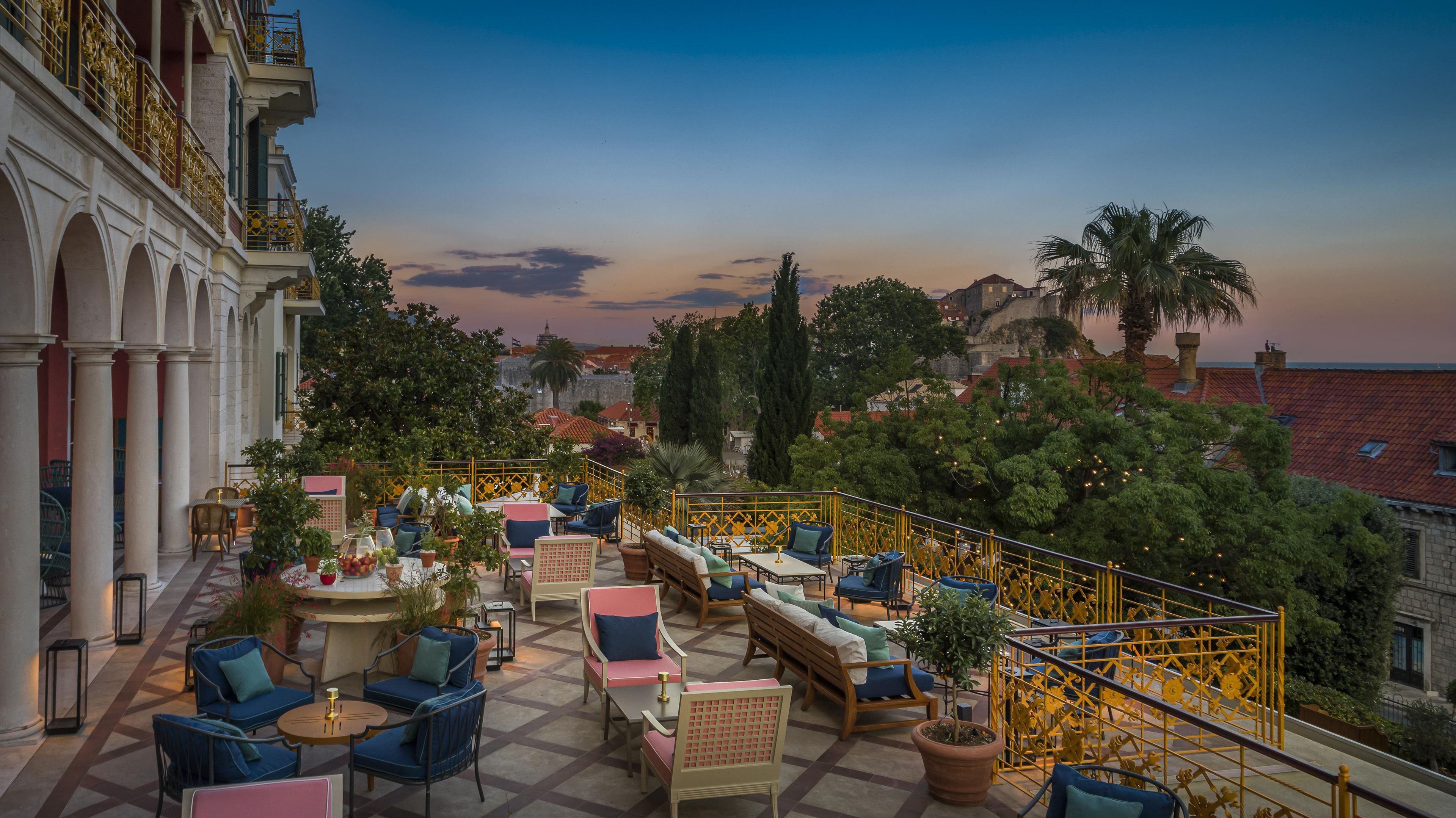 An outdoor seating area with tables and chairs, surrounded by greenery and overlooking a cityscape.