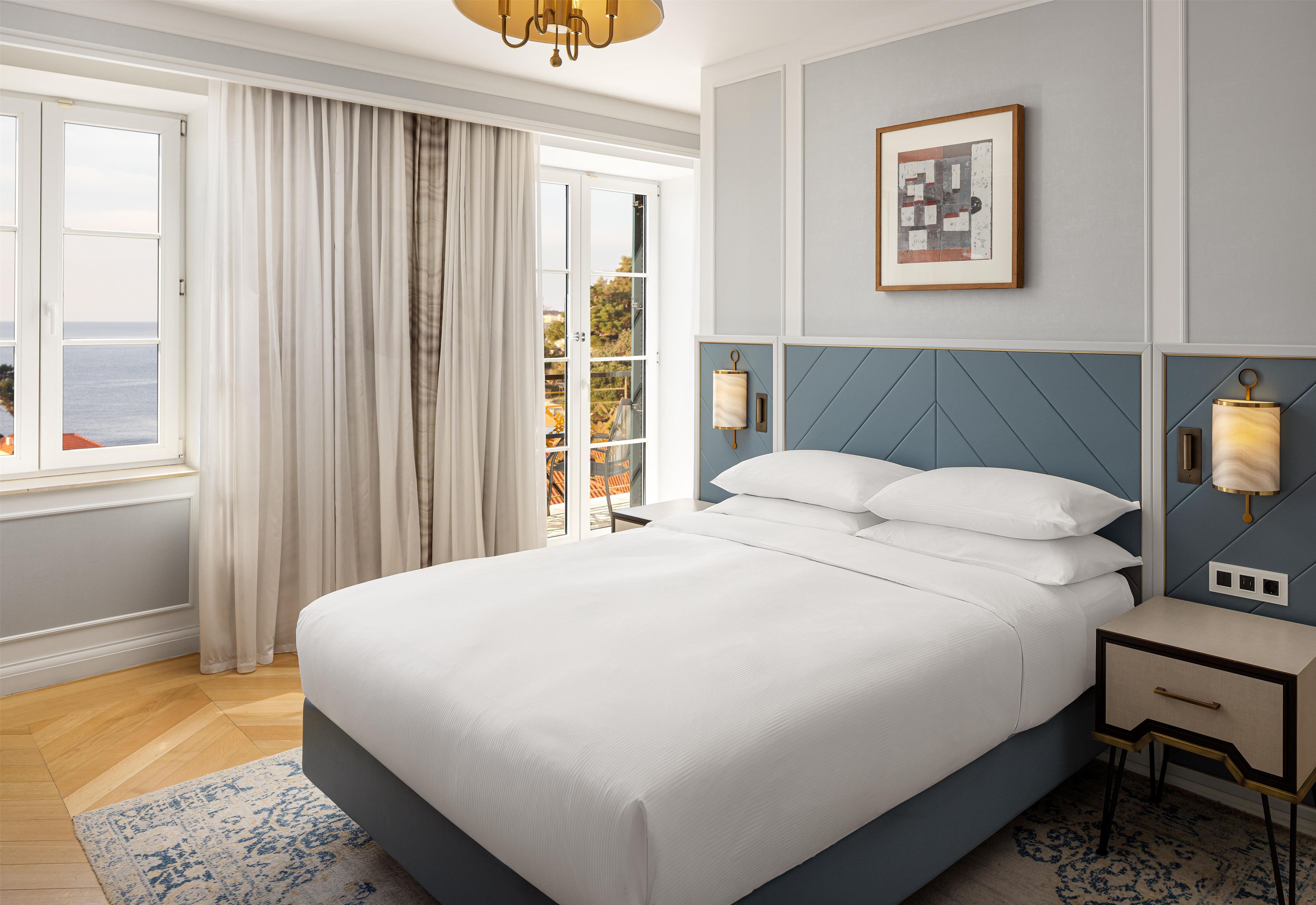 A hotel room with a large bed, a view of the sea through the window, a painting on the wall, and a nightstand with a lamp.