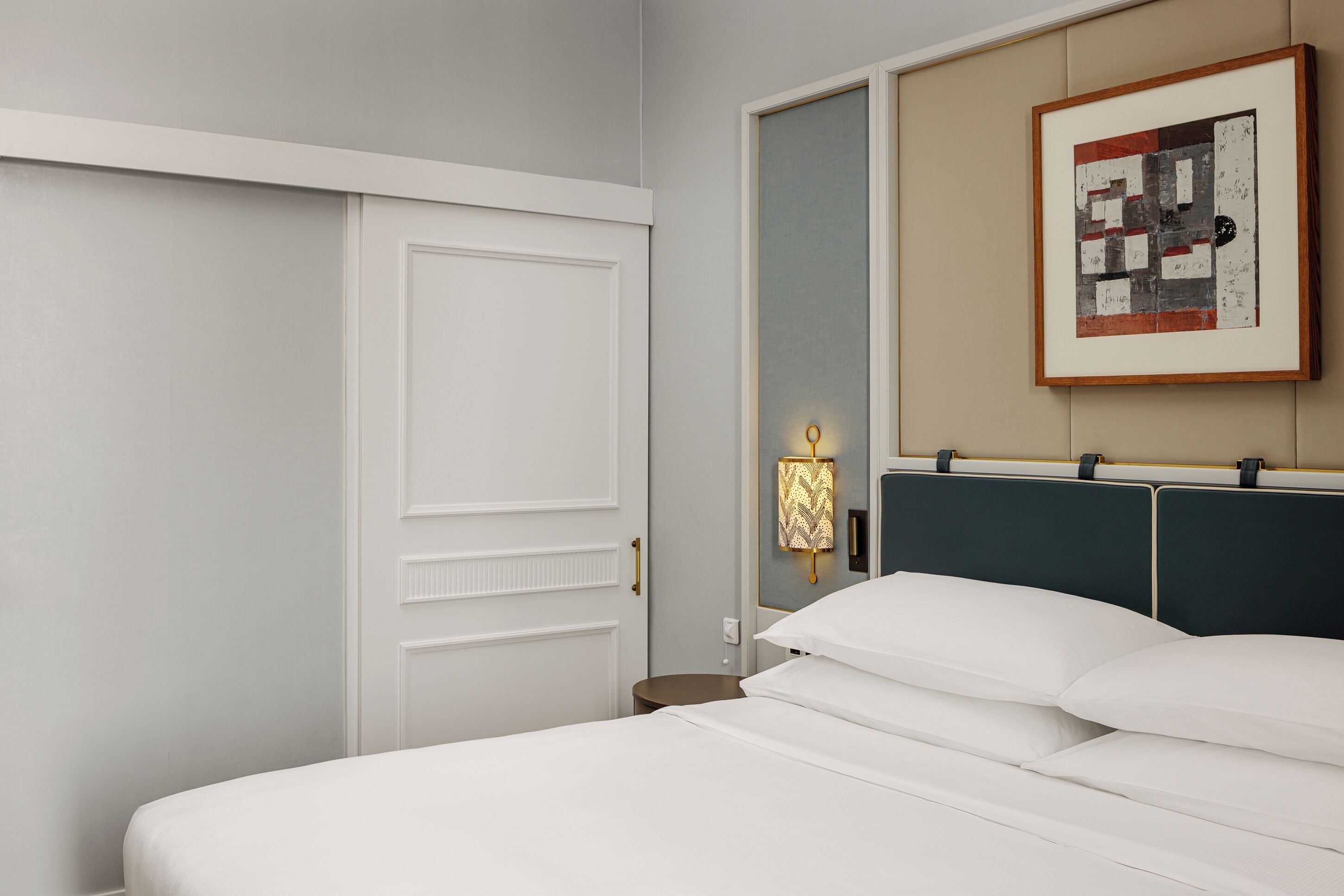 Quarto de hotel com cama, porta, luminária na parede e quadro emoldurado.