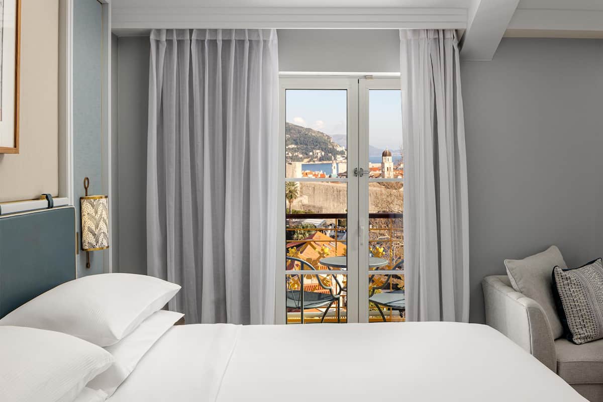 A hotel room with a bed, a sofa, a view of a town through a window, and a balcony with outdoor furniture.