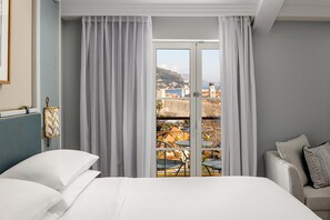 A hotel room with a bed, a sofa, a view of a town through a window, and a balcony with outdoor furniture. - Hilton Imperial Dubrovnik (Dubrovnik)