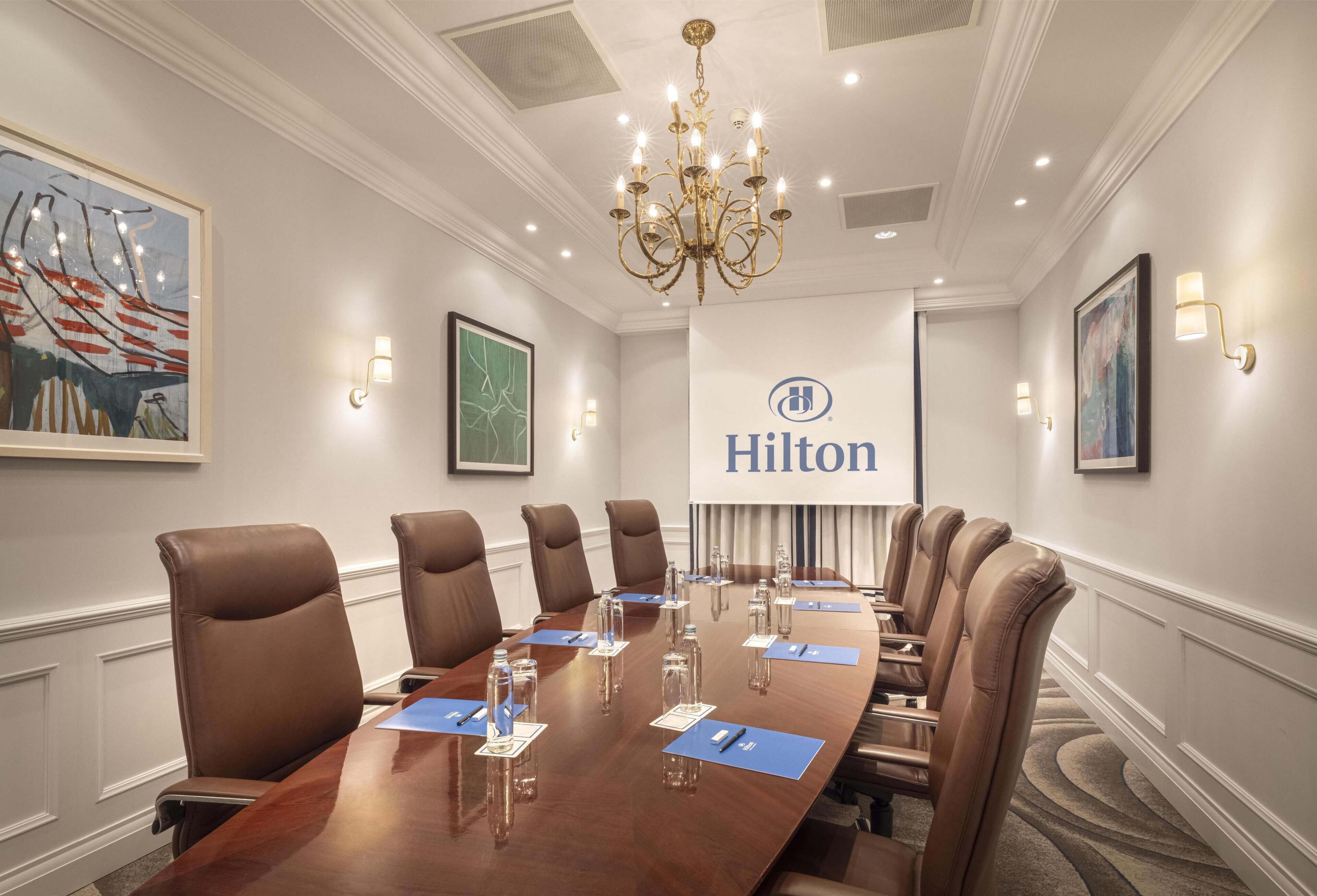 Un salón de conferencias con una larga mesa, sillas de cuero marrón, botellas de agua y el logotipo de Hilton en la pared.
