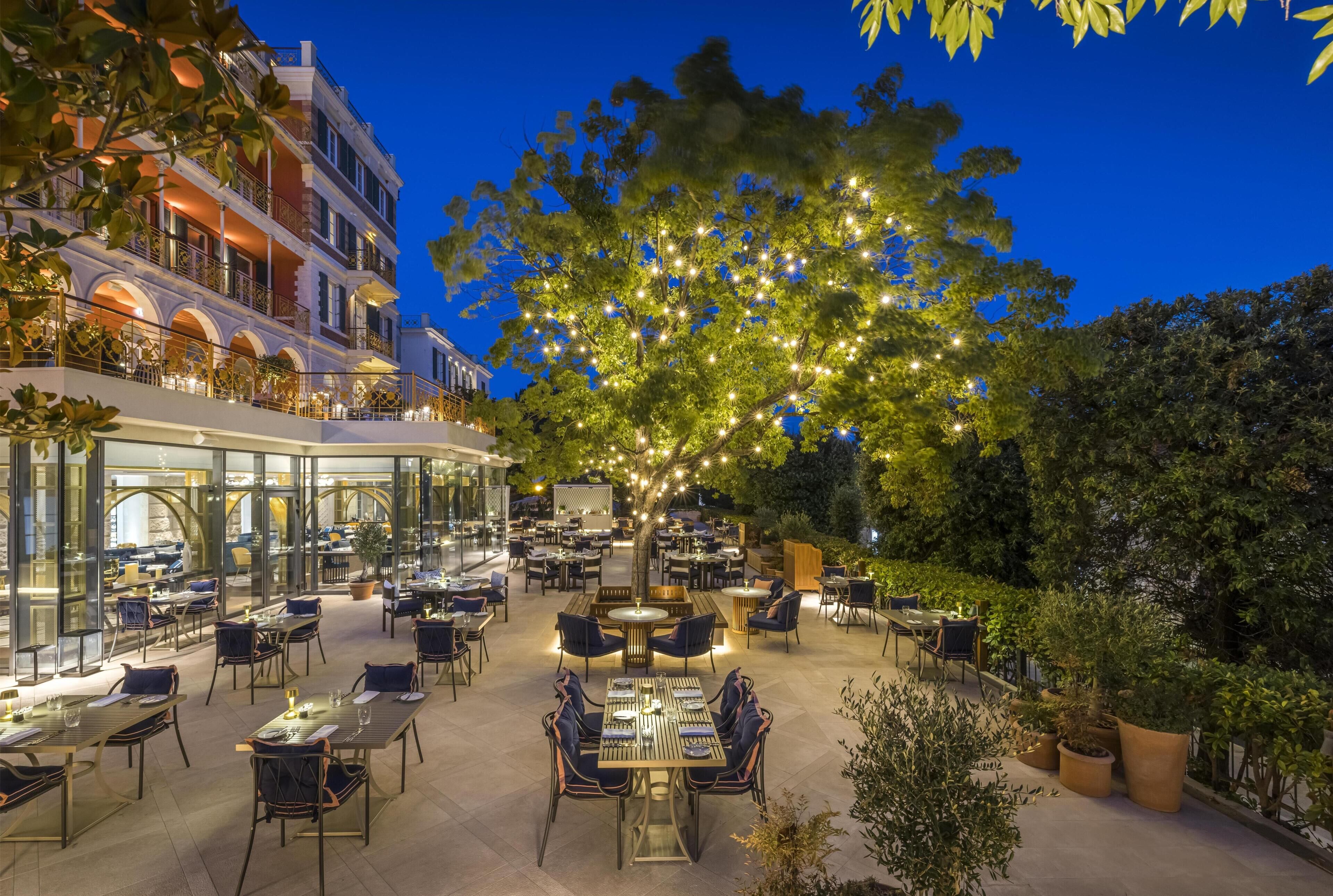 Un espacio de comedor al aire libre con mesas y sillas, un árbol grande con luces y un edificio con balcones.