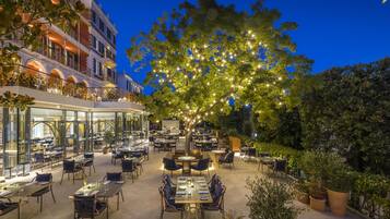 Un espacio de comedor al aire libre con mesas y sillas, un árbol grande con luces y un edificio con balcones.