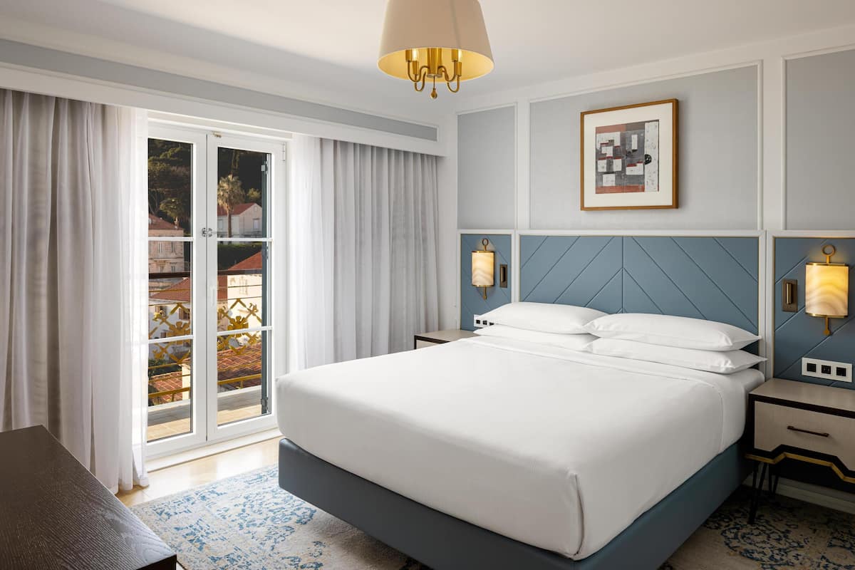 Een hotelkamer met een bed, nachtkastje, lamp, een lijst met afbeelding aan de muur en uitzicht door het raam.