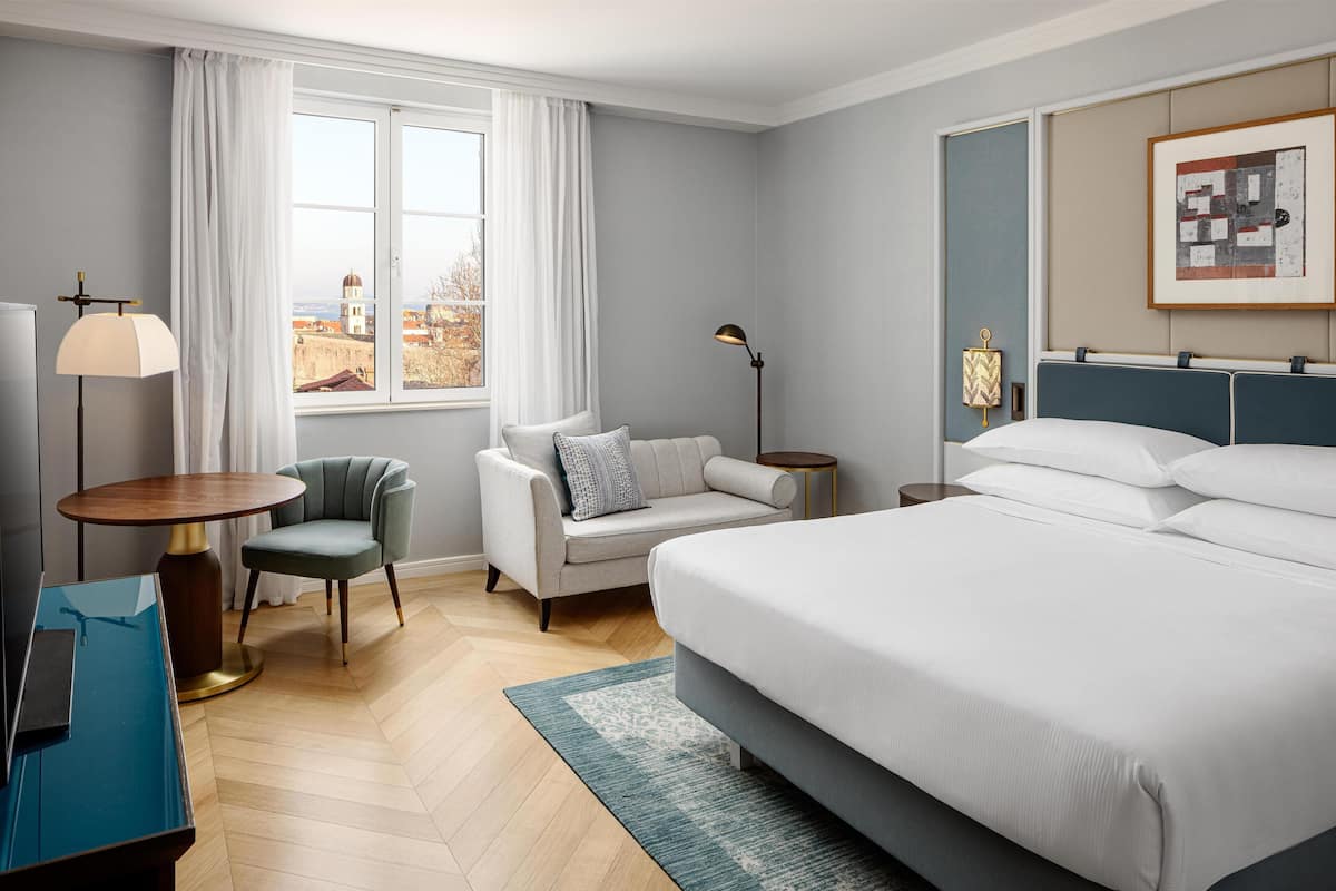 A hotel room with a bed, a chair, a desk, a lamp, and a window with a view.