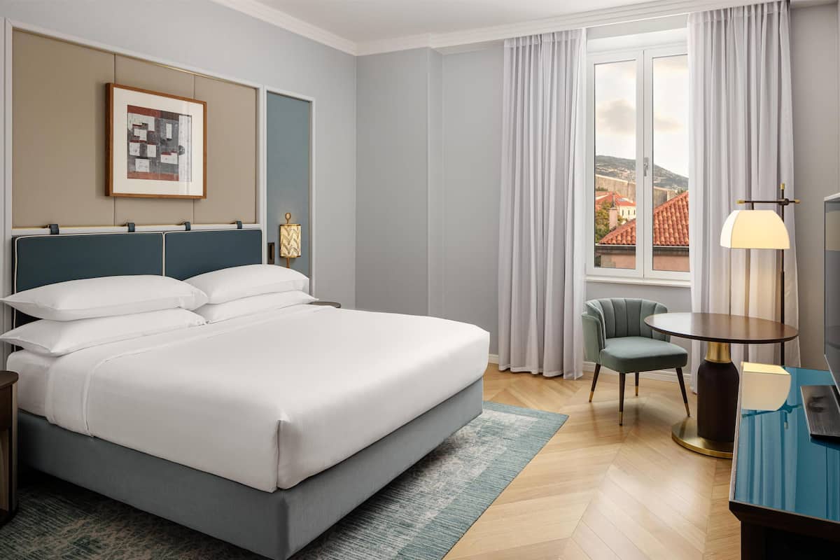Hotelkamer met een groot bed, een bureau, een stoel, een lamp, een inlijsting afbeelding aan de muur en een raam met gordijnen.