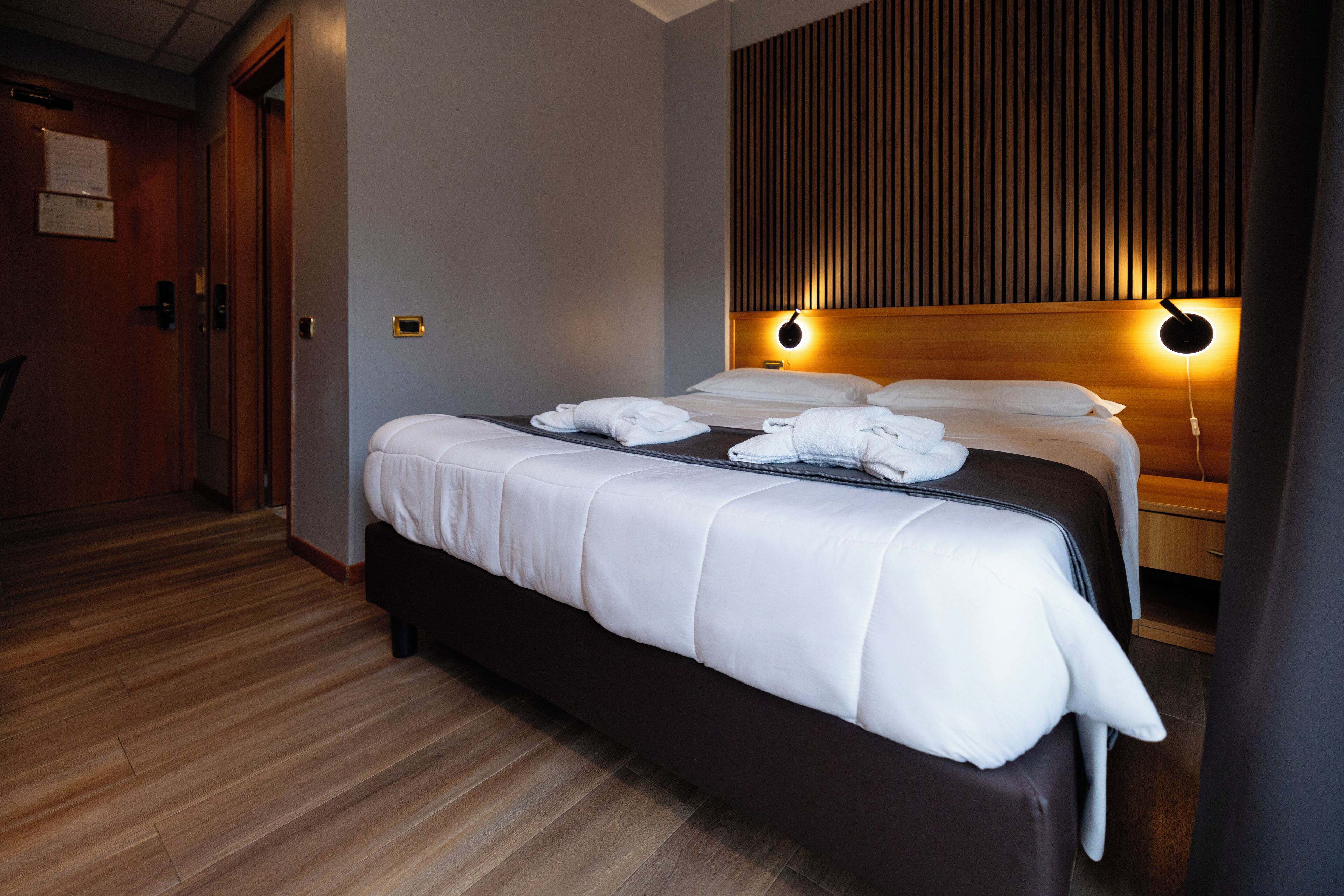 A hotel room with a large bed, wooden flooring, a wooden headboard, and two wall-mounted lights.