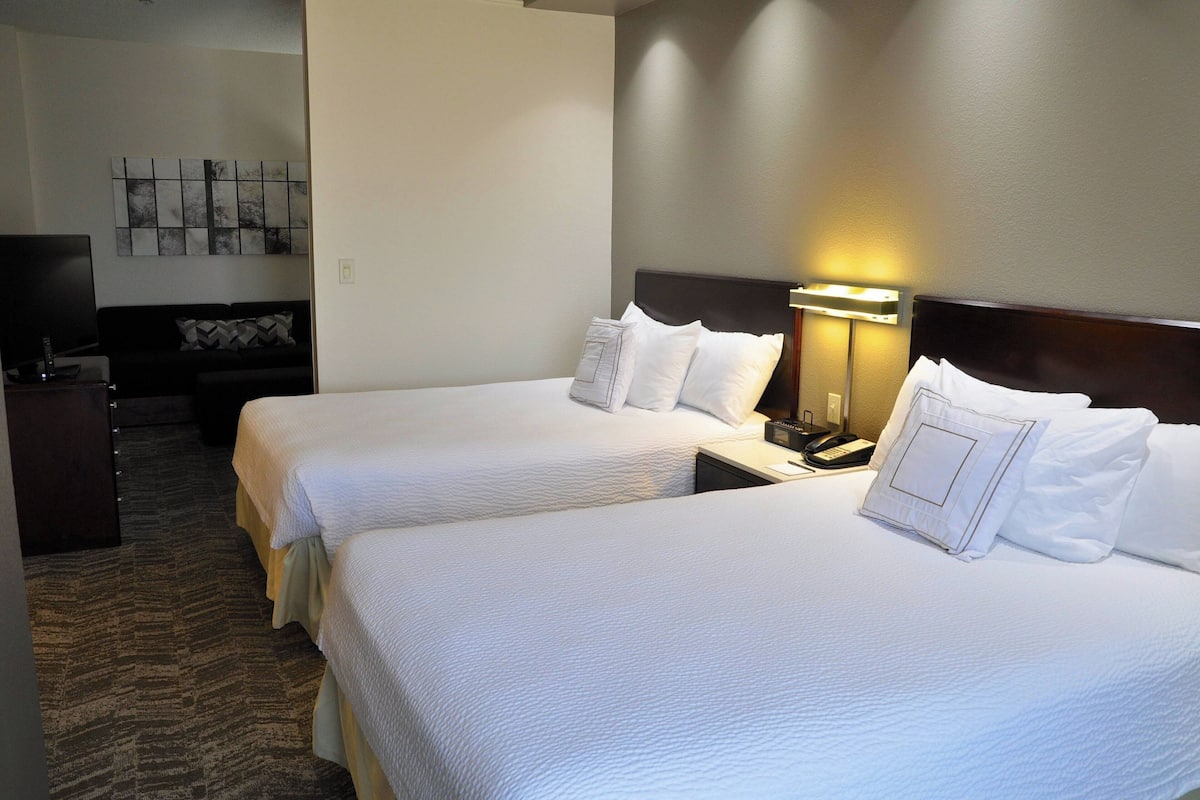 A hotel room with two beds, a desk with a lamp, a TV, and a wall art piece.