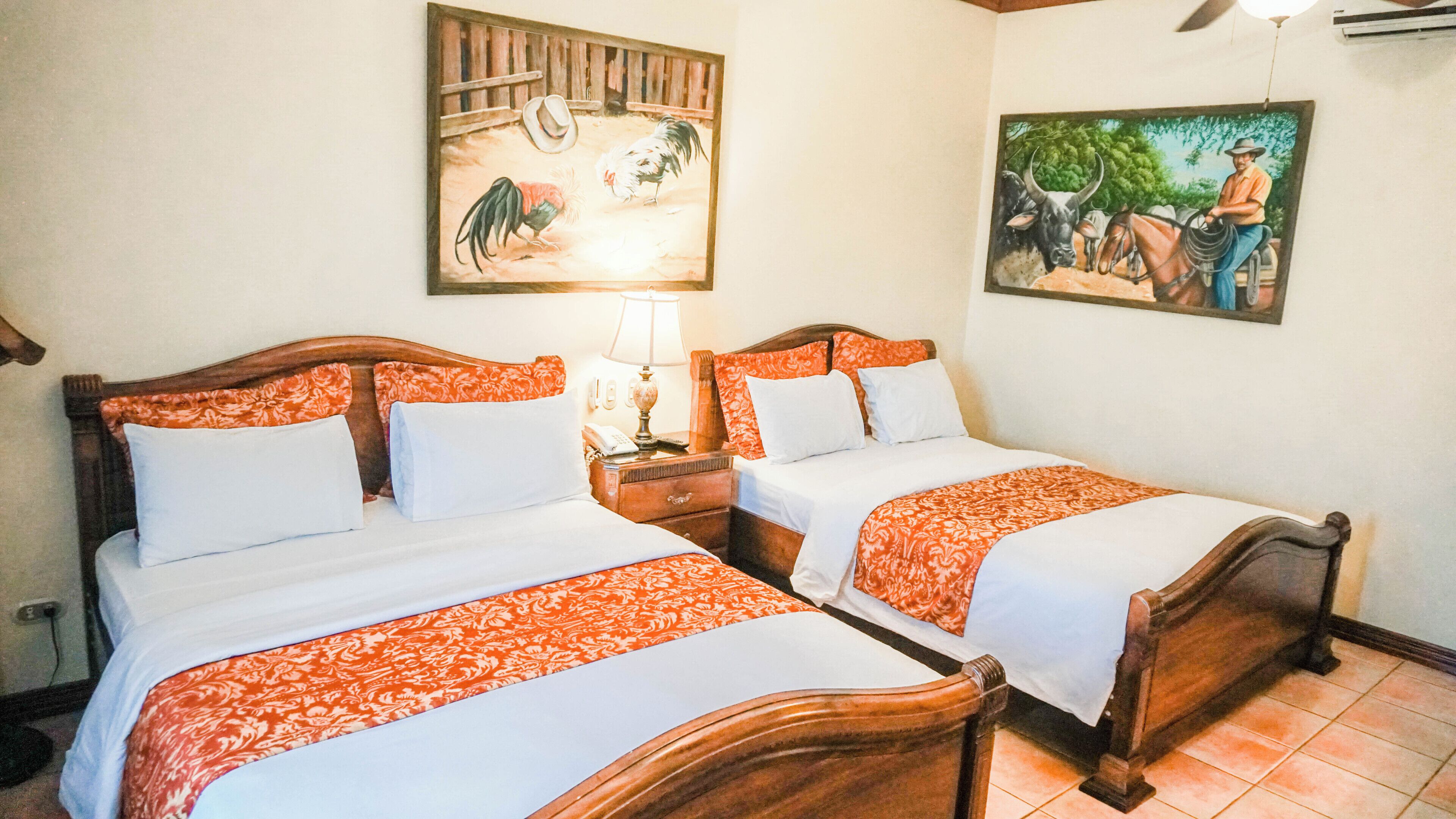 A hotel room with two beds, a nightstand, a lamp, and two paintings on the walls.