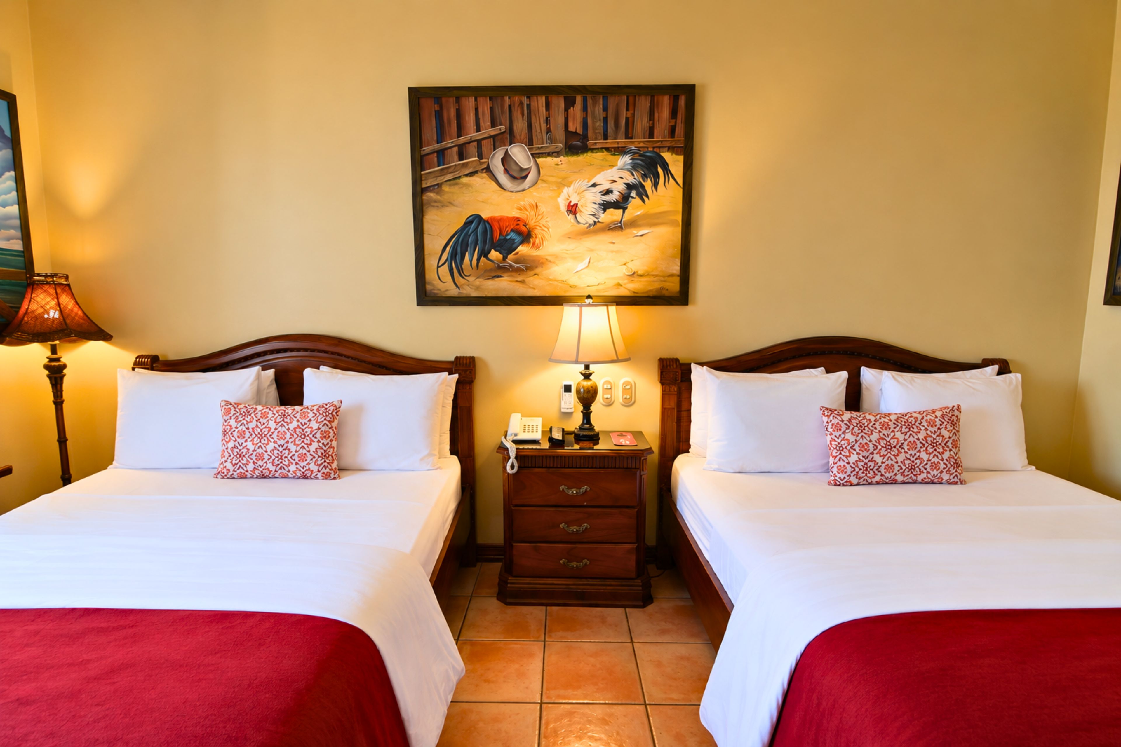 Habitación de hotel con dos camas, una mesita de noche, una lámpara, un cuadro de gallo y una colcha roja.