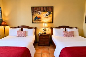 Habitación de hotel con dos camas, una mesita de noche, una lámpara, un cuadro de gallo y una colcha roja.