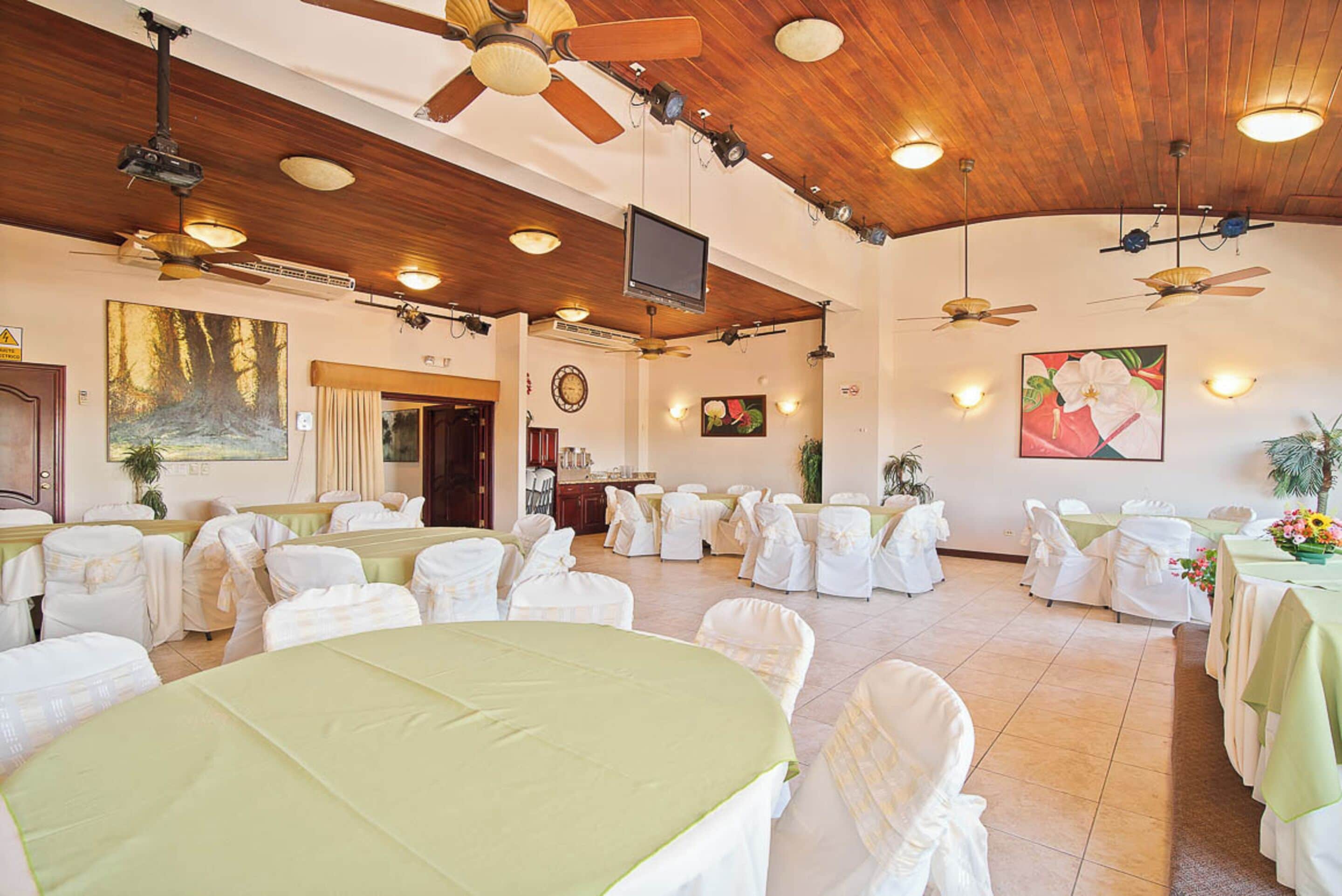 A banquet hall with round tables covered in white tablecloths and chairs, wooden ceiling, and ceiling fans.