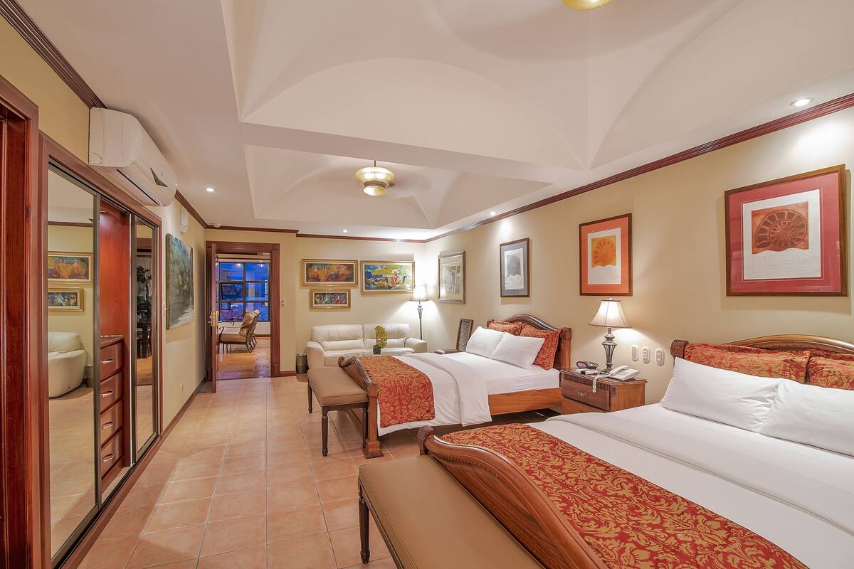 A hotel room with two beds, a desk, a chair, a bench, and a door leading to another room.