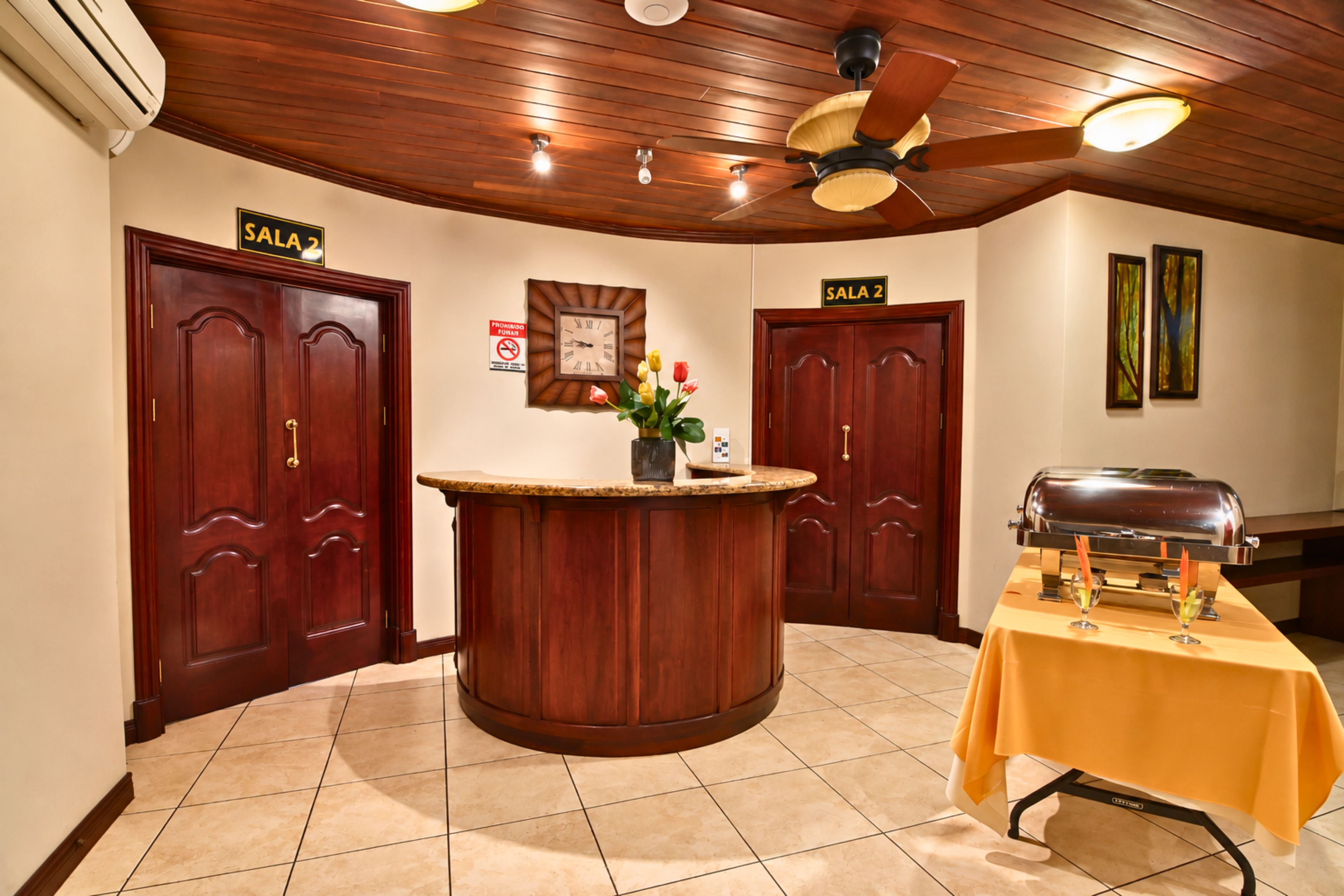 A lobby with two wooden doors labeled 'SALA 2', a reception desk, a clock, a 'No Smoking' sign, and a grill table.