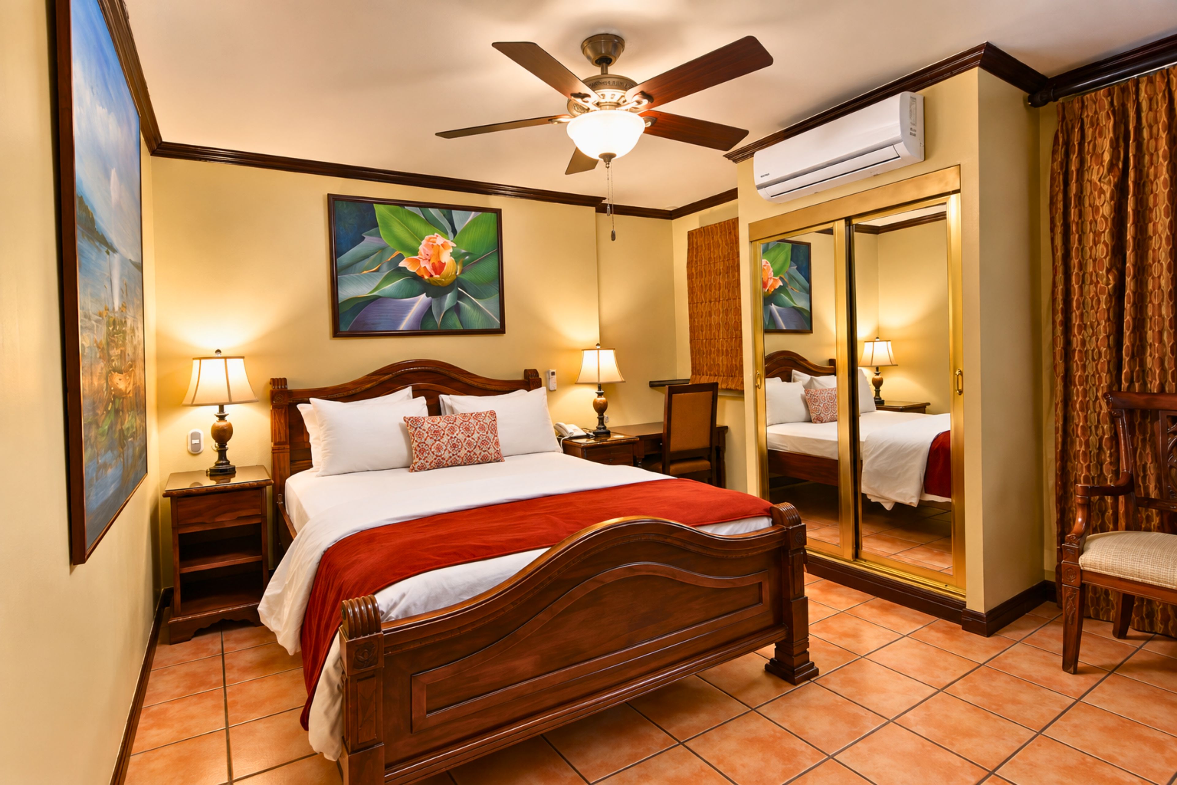 A hotel room with a wooden bed, two bedside lamps, a ceiling fan, a large mirror, and a painting on the wall.