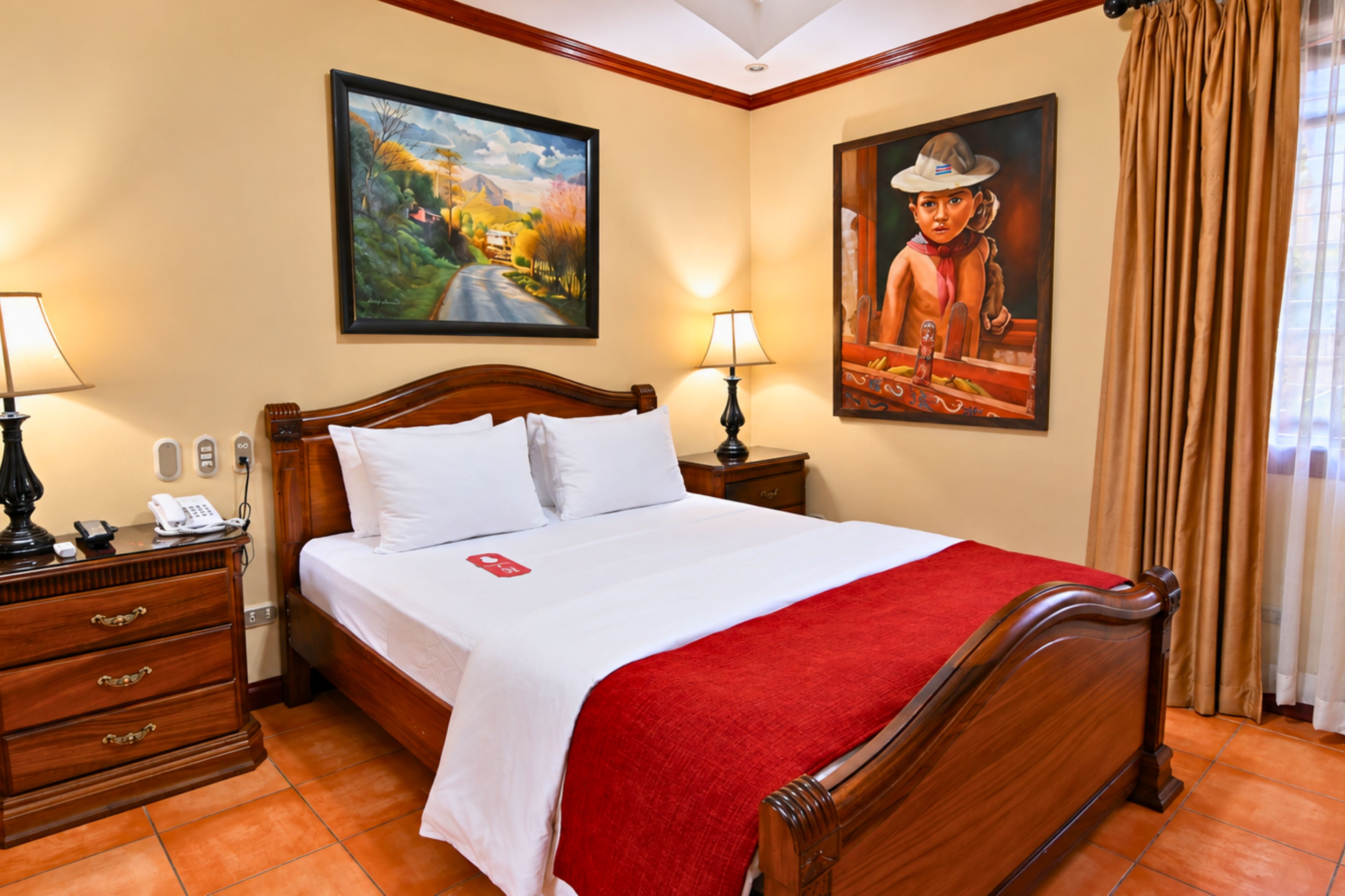 Habitación de hotel con cama de madera, una manta roja, dos lámparas de noche, un teléfono, una mesita de noche y dos cuadros en las paredes.