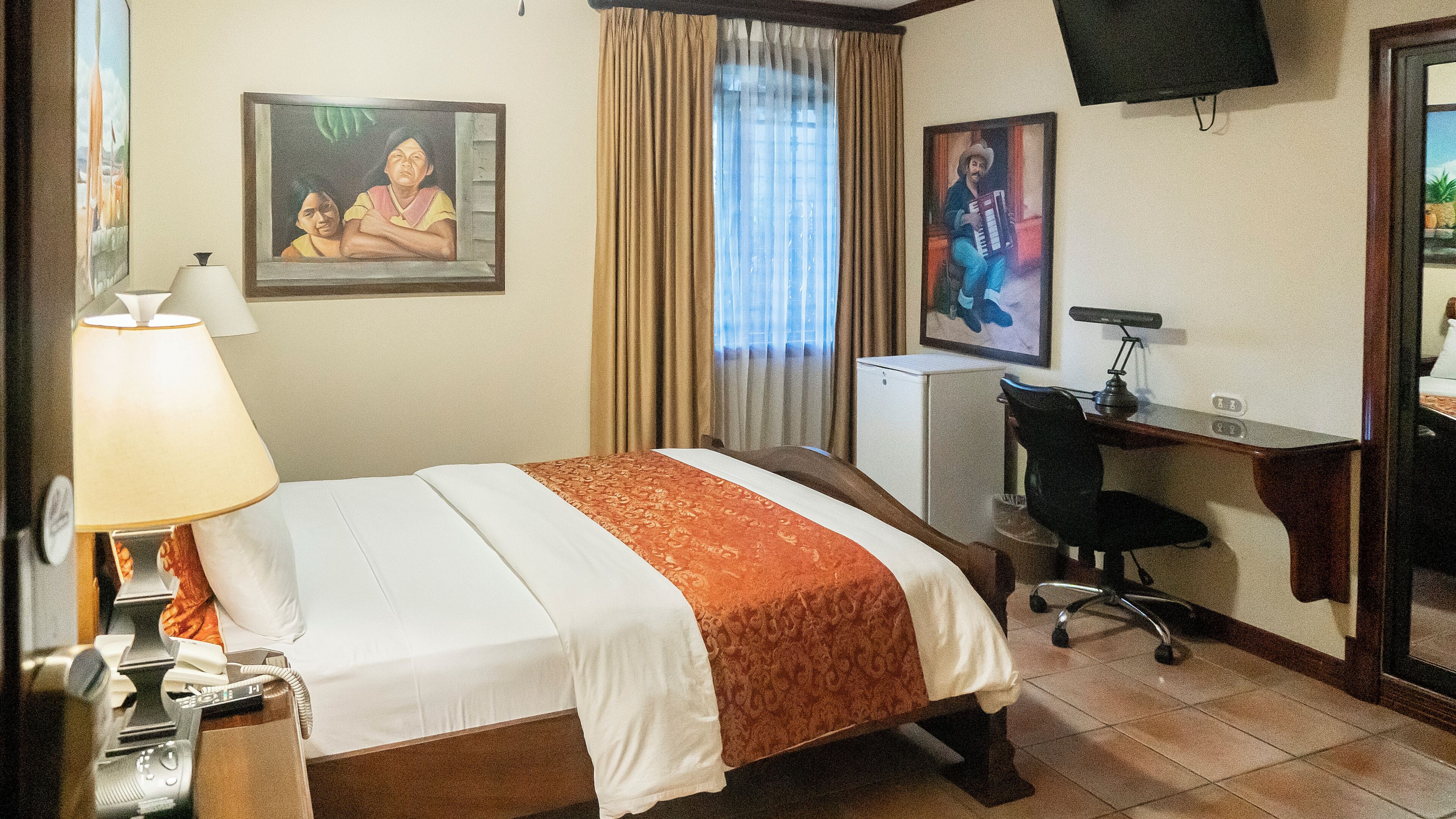 Une chambre d’hôtel avec un lit, un bureau, une chaise, une lampe et deux tableaux au mur.