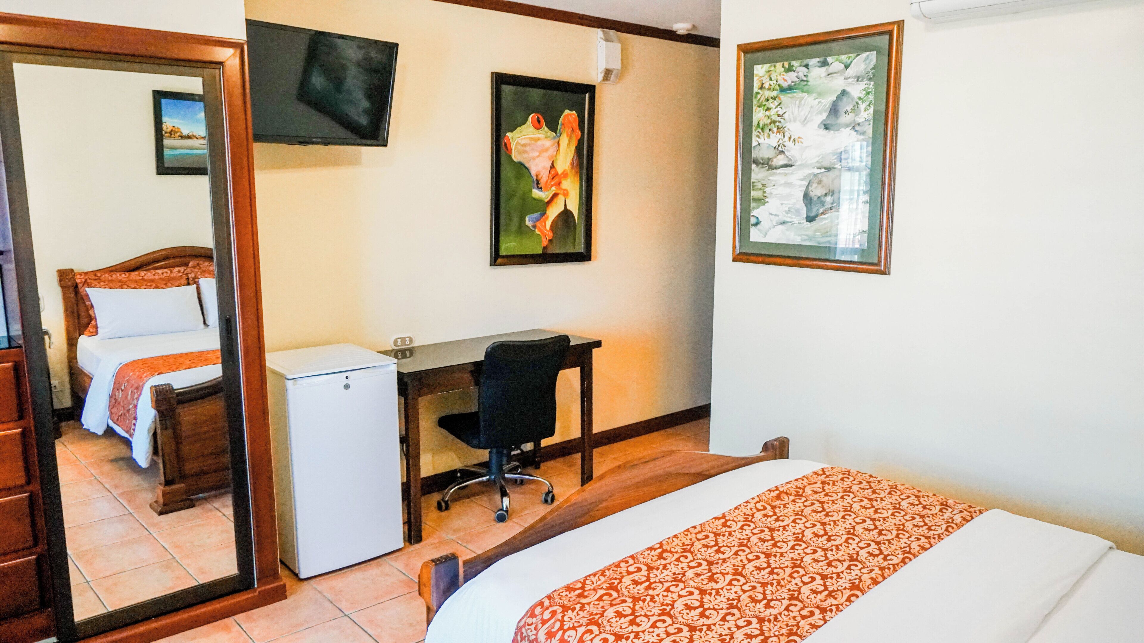 A hotel room with a bed, a desk, a chair, a small refrigerator, a mirror, and two framed pictures on the wall.