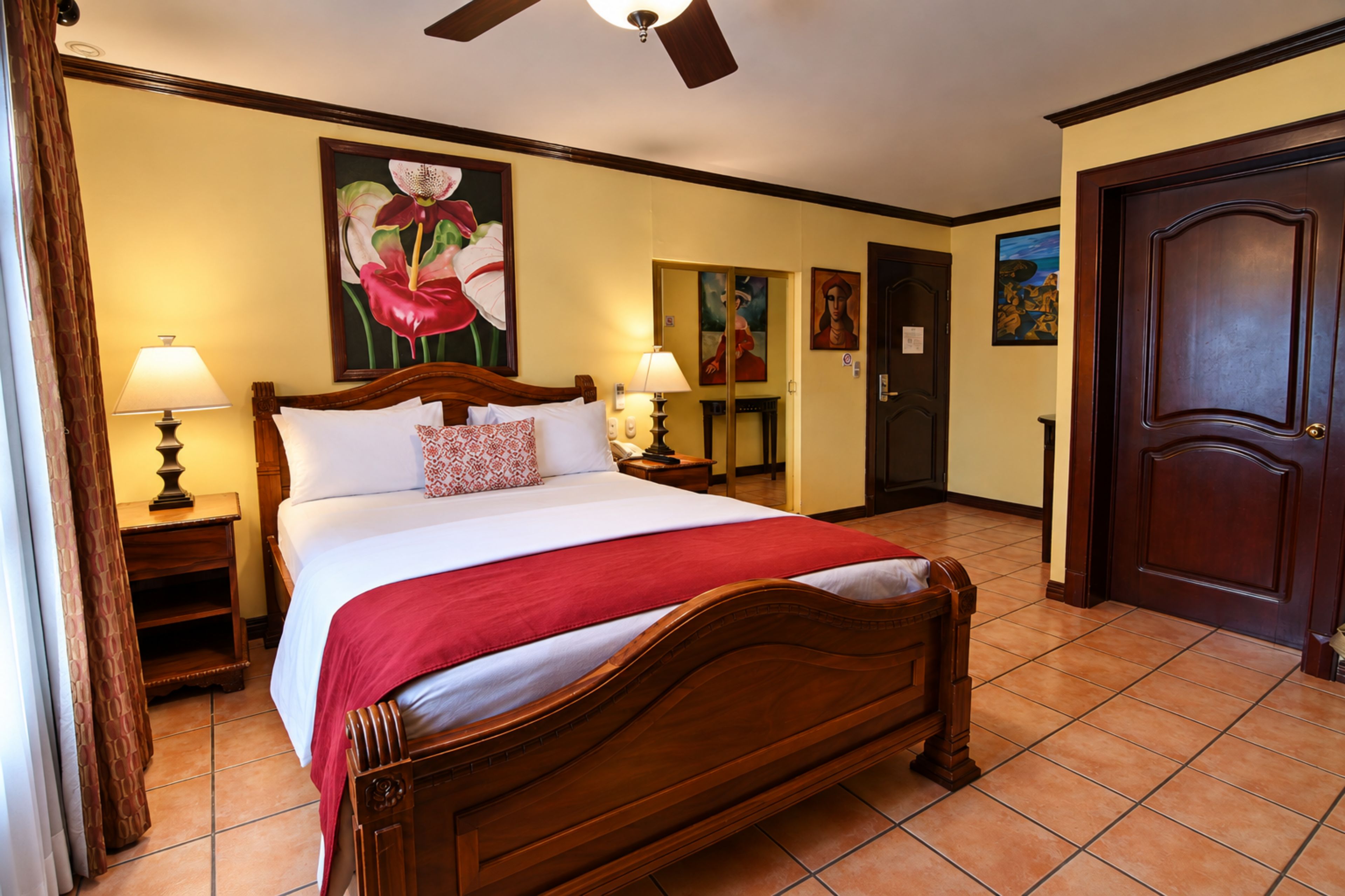 A hotel room with a wooden bed, two bedside lamps, a red throw blanket, and artwork on the walls.