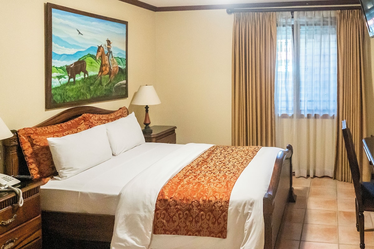 A hotel room with a bed, two bedside lamps, a telephone, a painting, a window with curtains, and a chair.