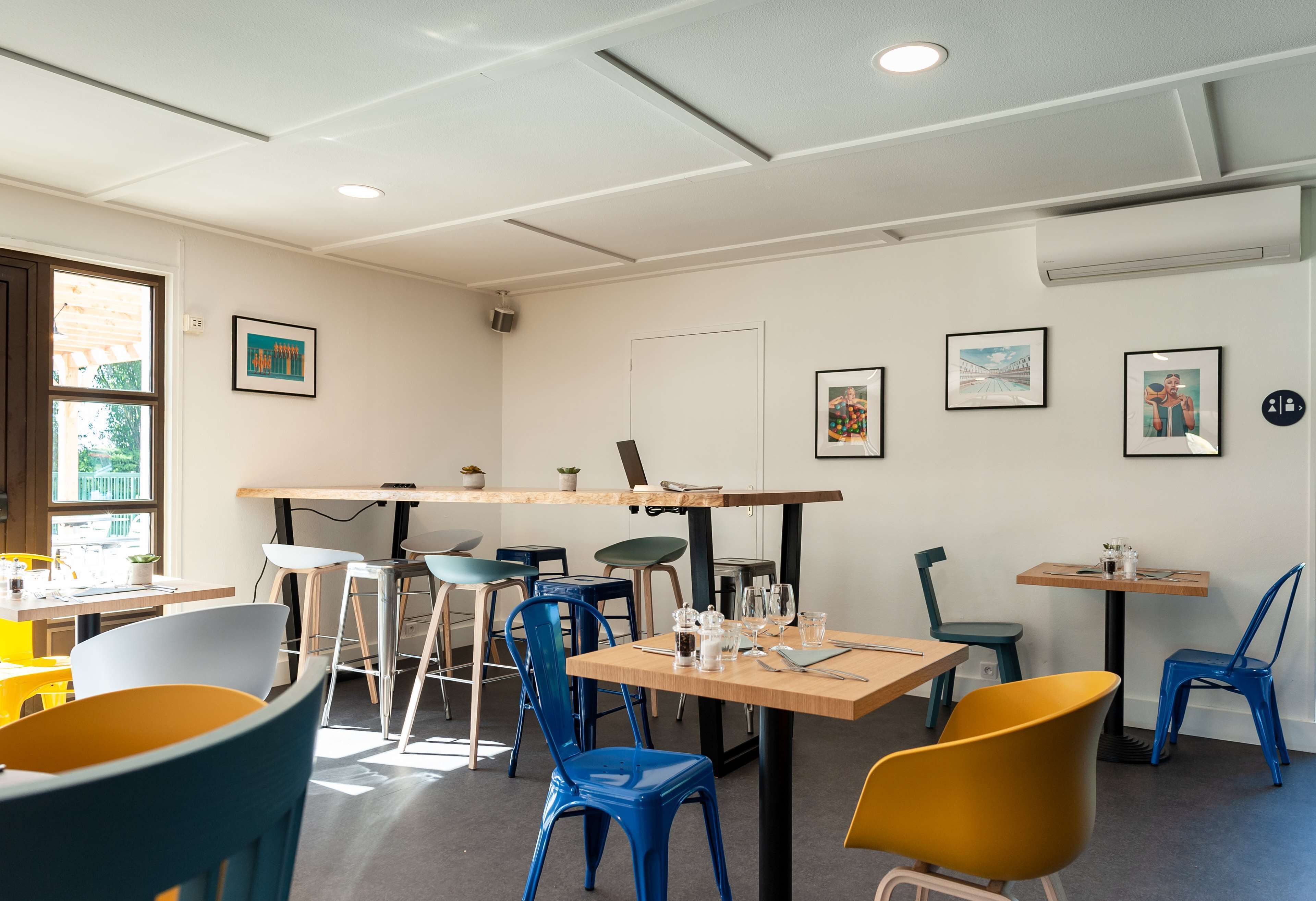 A modern dining area with multiple tables and chairs, framed artwork on the walls, and a window providing natural light.