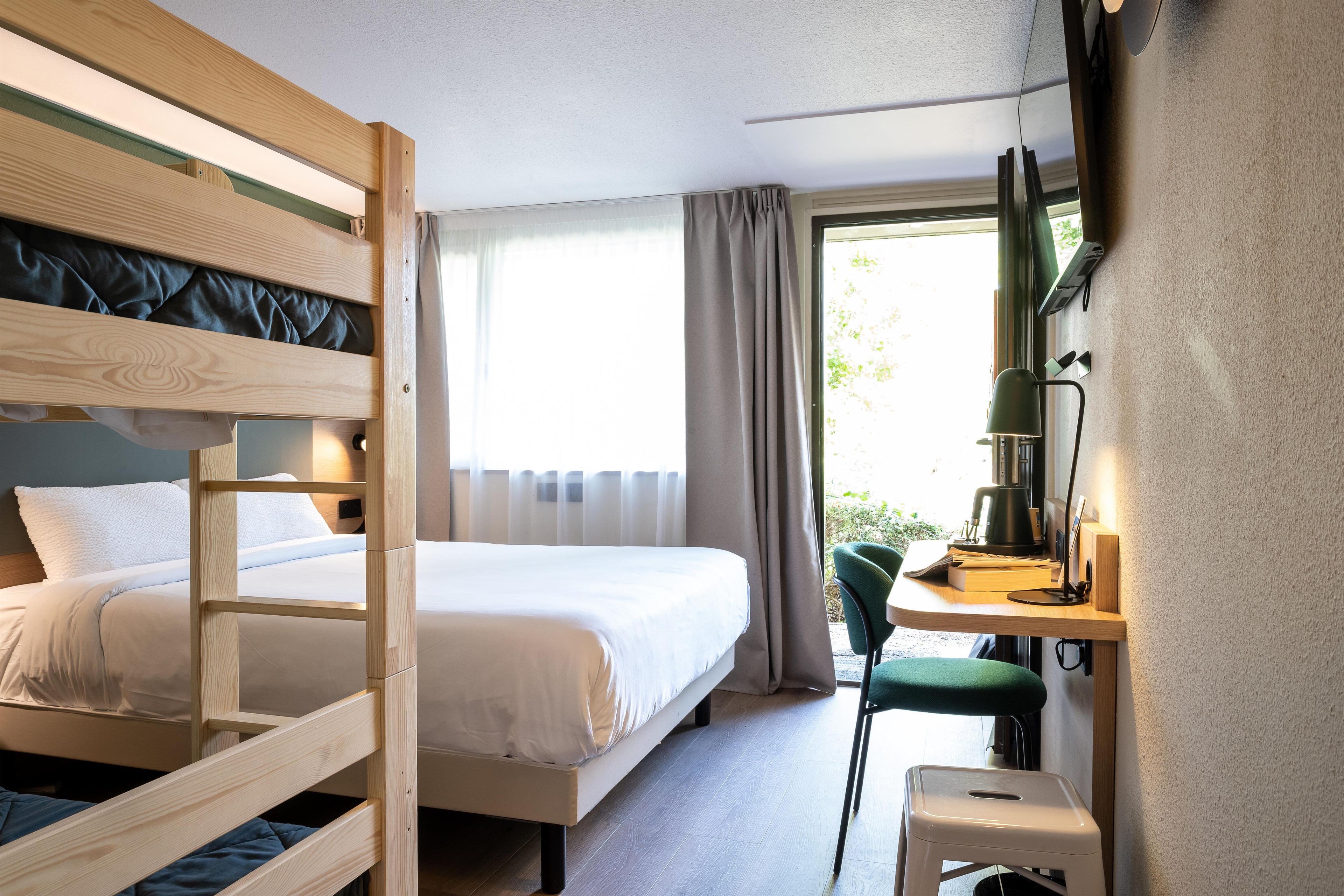 Een hotelkamer met een stapelbed, een bureau met een stoel, een raam met gordijnen en uitzicht op groen.