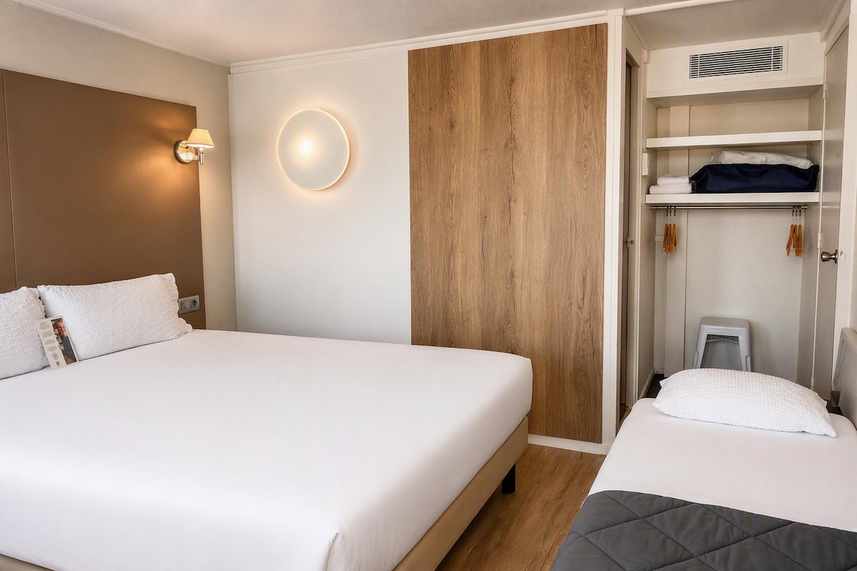 A hotel room with a large bed, a smaller bed, a wardrobe with shelves, and a bedside table.