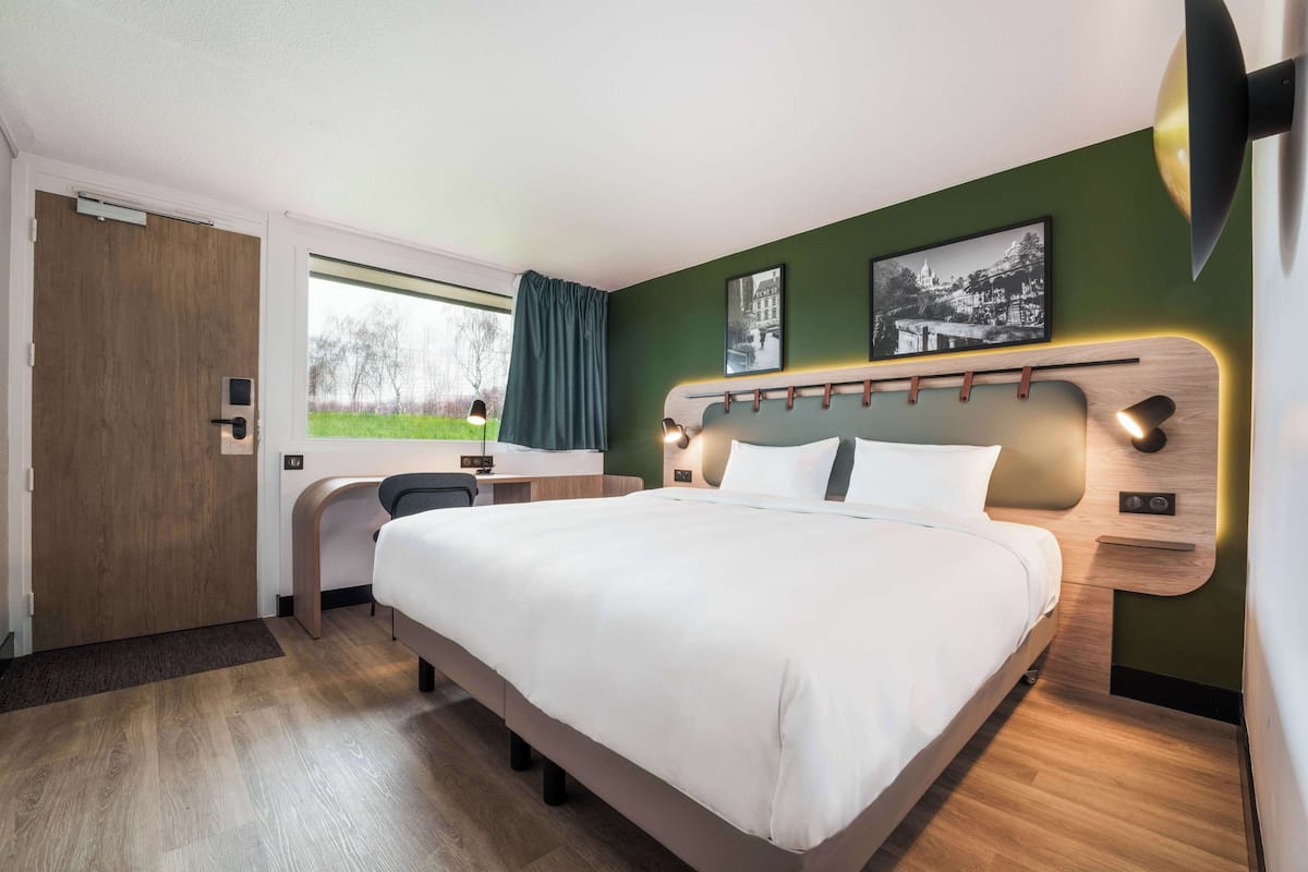 A modern hotel room with a large bed, a desk, and a chair. There is a window with a view of greenery outside.