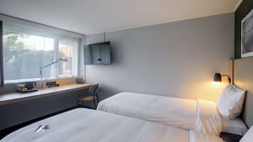 A hotel room with two beds, a desk with a chair, a TV mounted on the wall, and a window with a view of trees.
