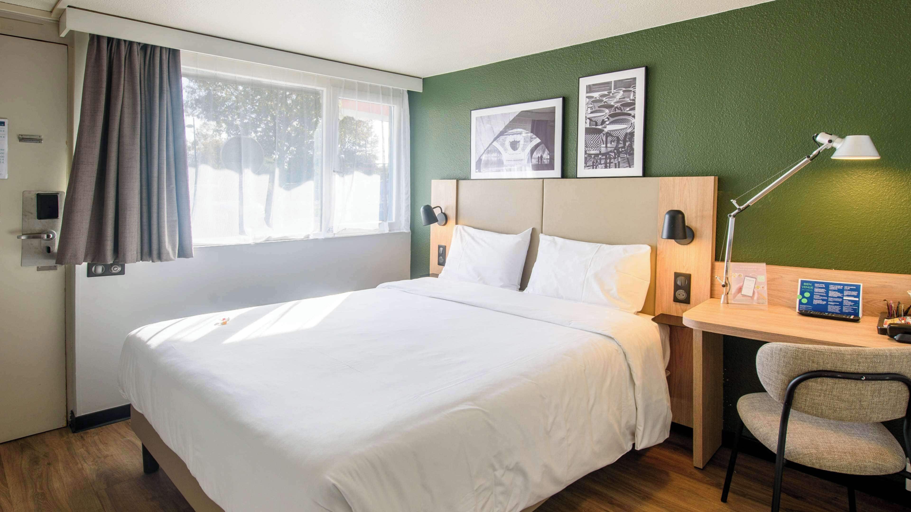 A hotel room with a bed, a desk with a chair, a lamp, and two framed pictures on the wall.