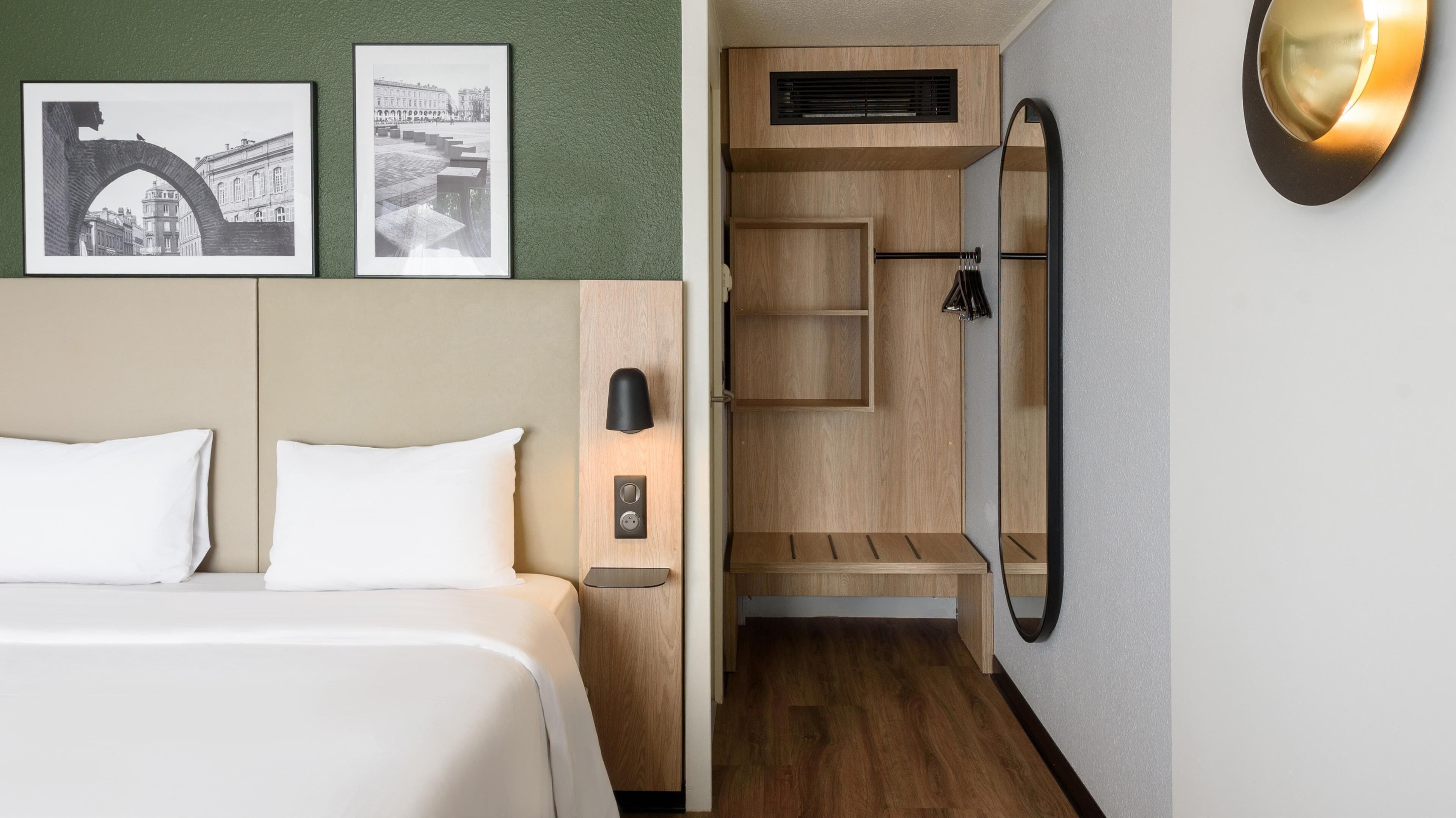 A hotel room with a bed, a nightstand, a wall-mounted lamp, and a wooden shelving unit.
