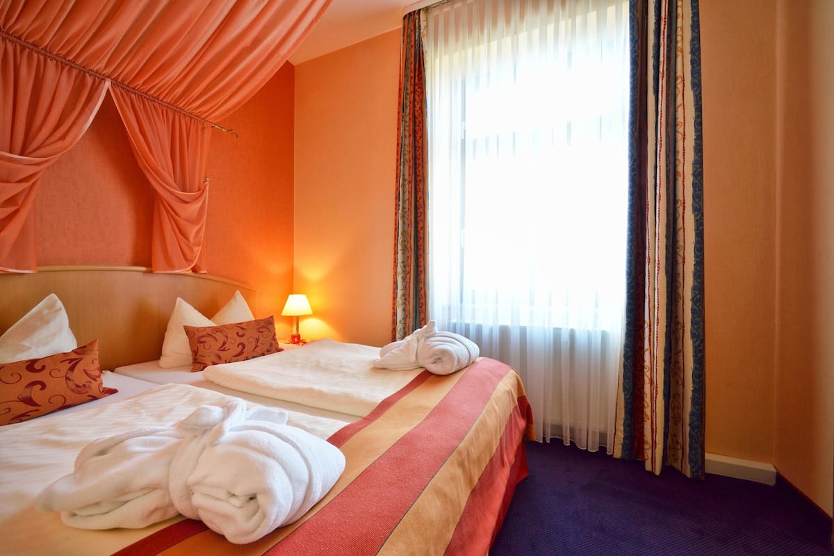 A hotel room with two beds, a large window with curtains, and a lamp on the nightstand.