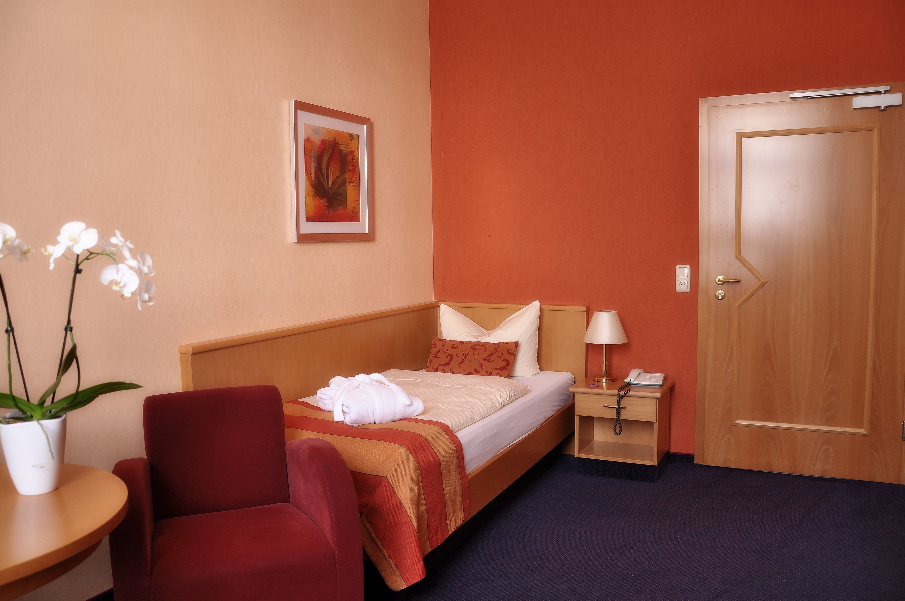 Una habitación de hotel con cama, una silla, mesita de noche con lámpara, teléfono y puerta.