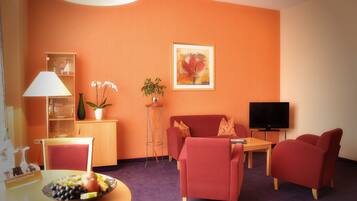 Een woonkamer met een bank, fauteuils, een televisie, een salontafel, een schaal met fruit, een lamp, een plant en een schilderij aan de muur.