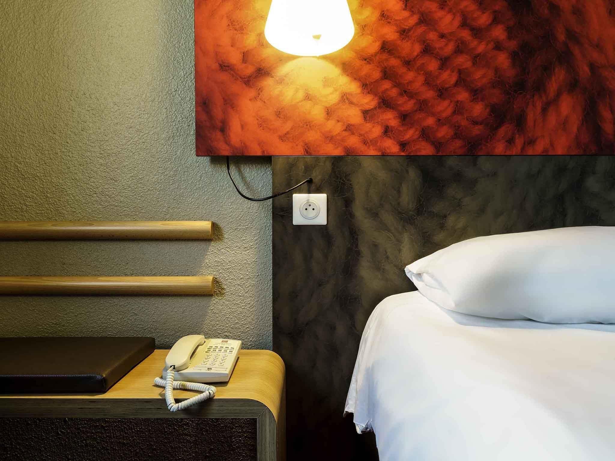 A hotel room with a bed, a bedside telephone, and a wall-mounted light fixture.