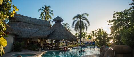A pool area with thatched-roof structures, palm trees, and a clear sky.
