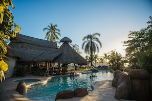 A pool area with thatched-roof structures, palm trees, and a clear sky.