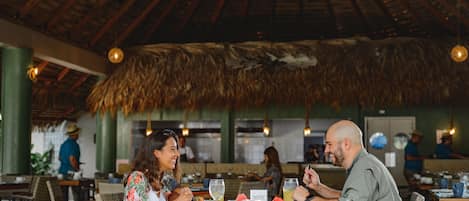 A restaurant with wooden tables and chairs, a thatched roof, and hanging lights.